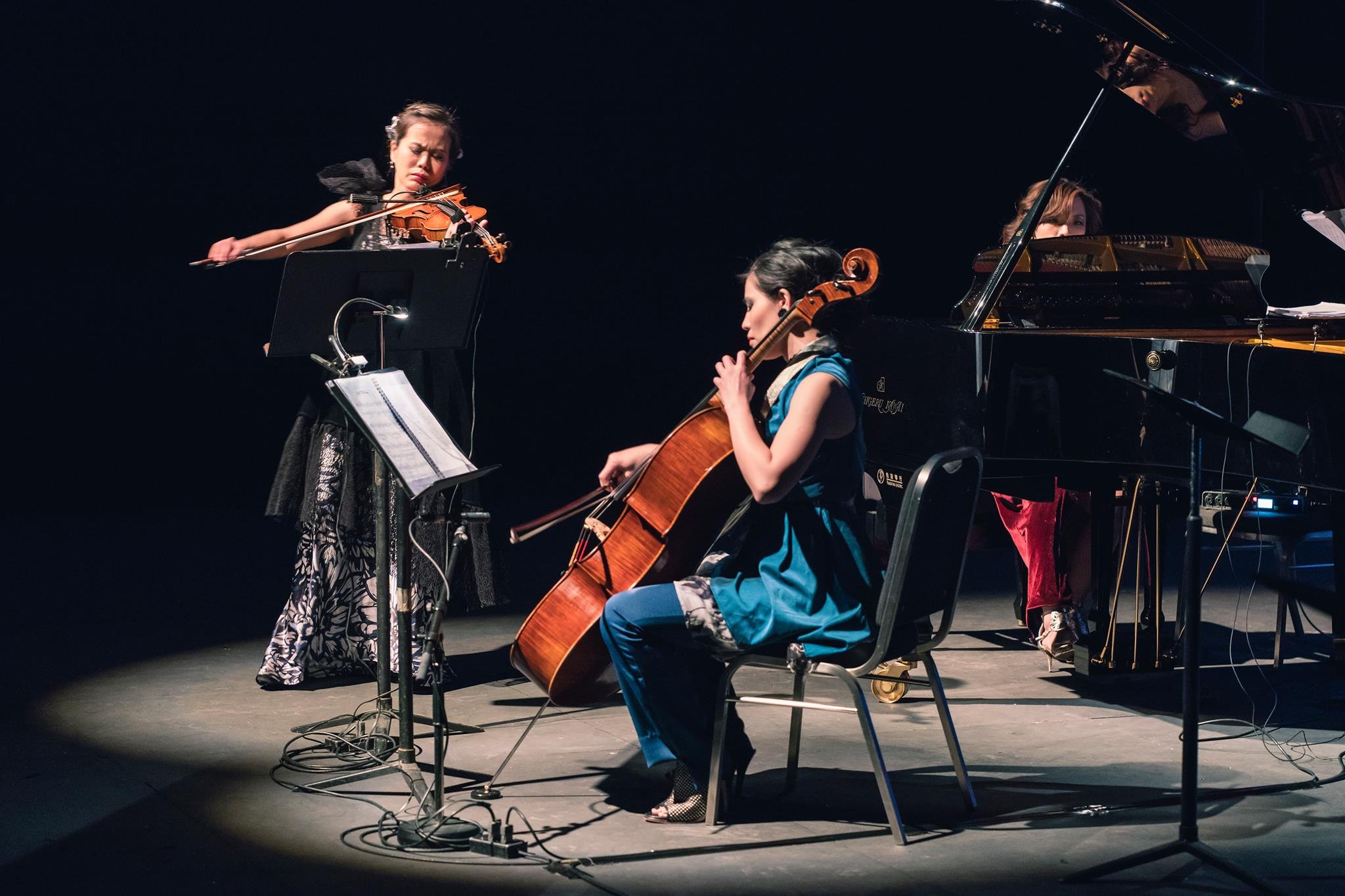 Three women are performing music on stage. One is playing a violin, another a cello, and the third a grand piano.