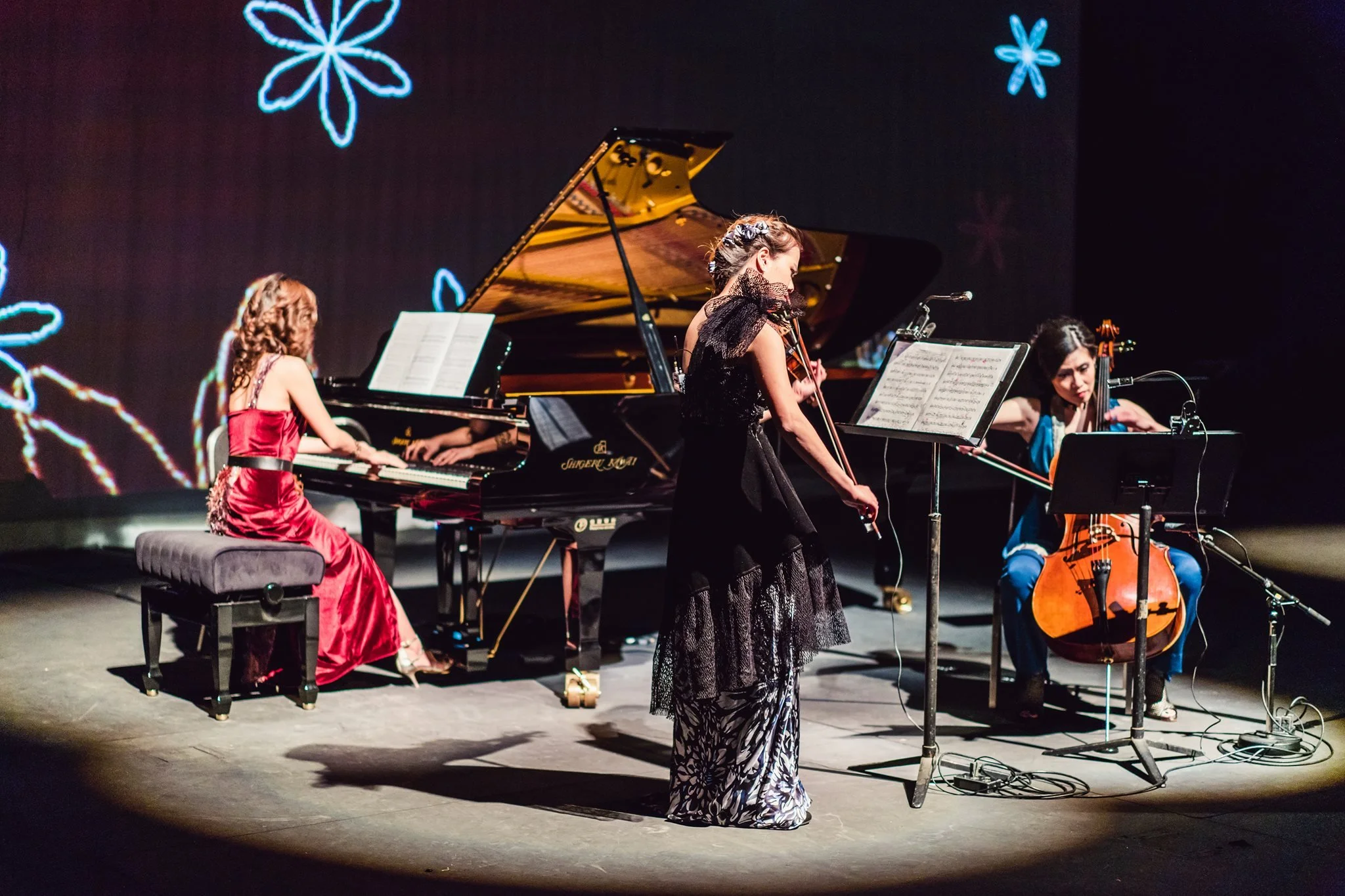 A group of four female musicians performing on a stage. One playing the grand piano, another playing the violin, and the third playing the cello. A woman in a black dress is standing with a microphone in front of them. The background has a black curt