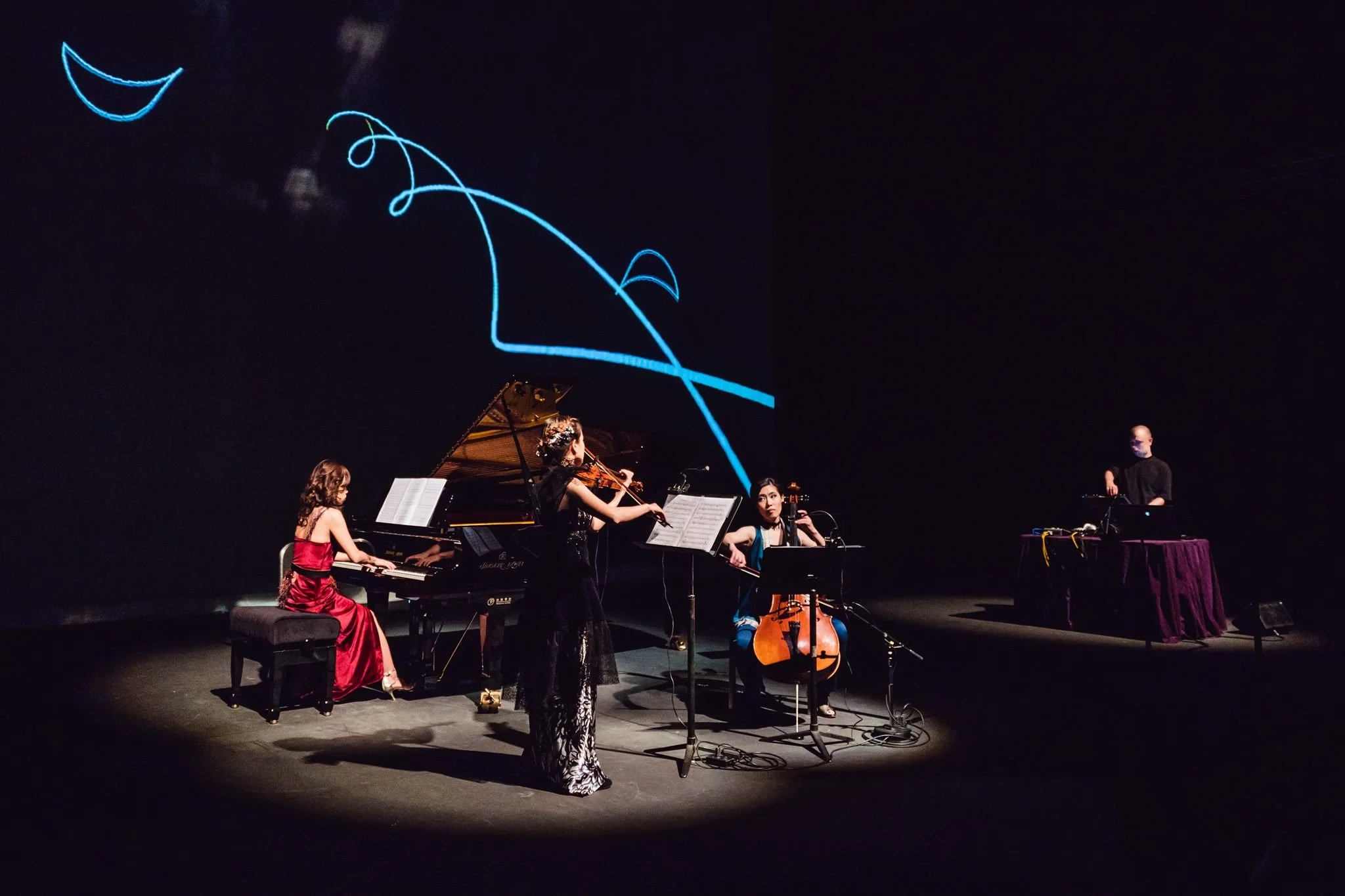 A group of four women and one man performing on stage with musical instruments, including a grand piano, a cello, a violin, and a keyboard. A dark background features abstract, glowing blue line art of a crescent moon and swirling shapes.