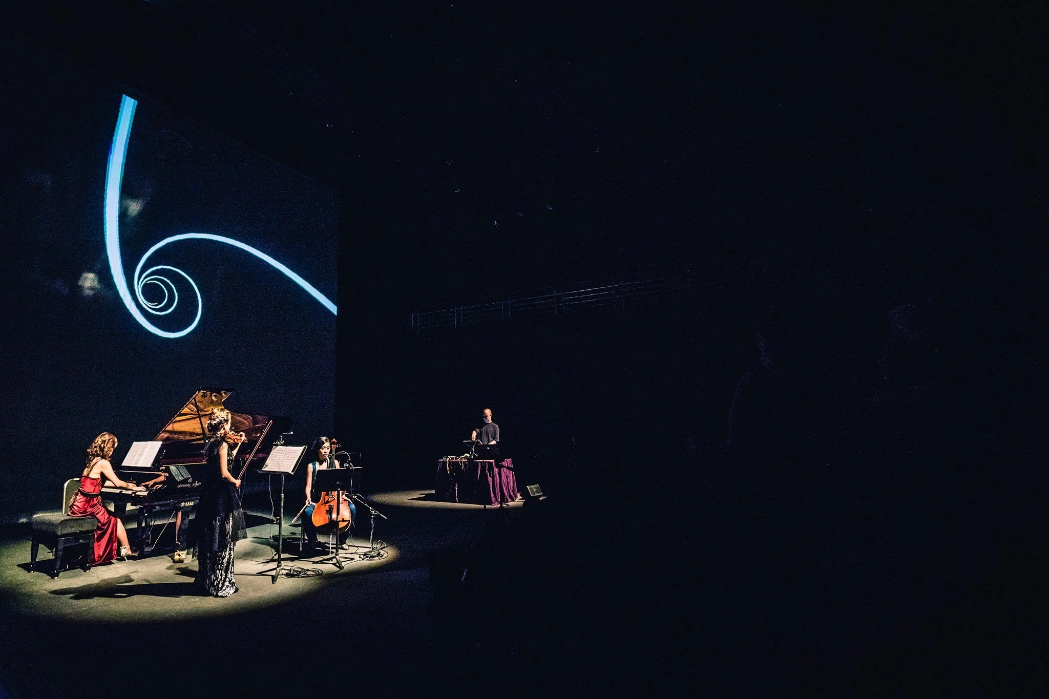 A small orchestra performing on stage under a starry night sky projection, with a spiral galaxy design on the backdrop, and a musician in a red dress playing the piano.