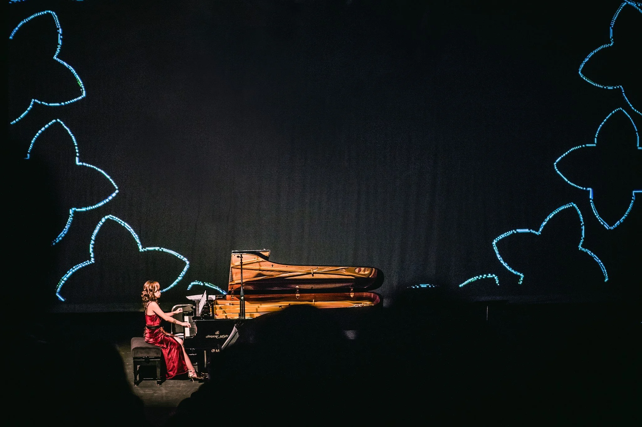 A young girl in a red dress playing a grand piano on a stage with a dark background and blue neon-like designs.