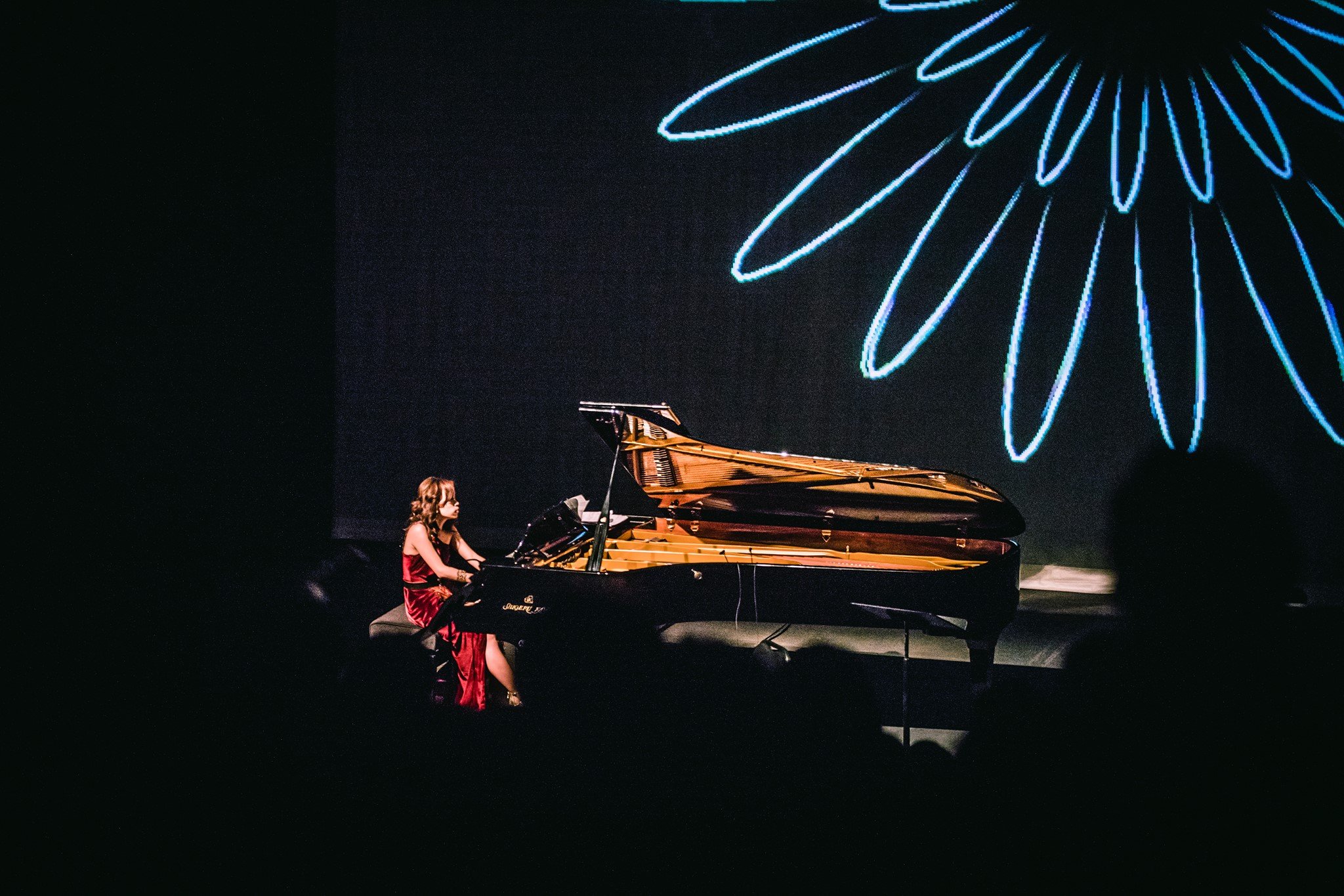 A woman in a red dress playing a grand piano on a stage with a backdrop of a large, illuminated flower design.