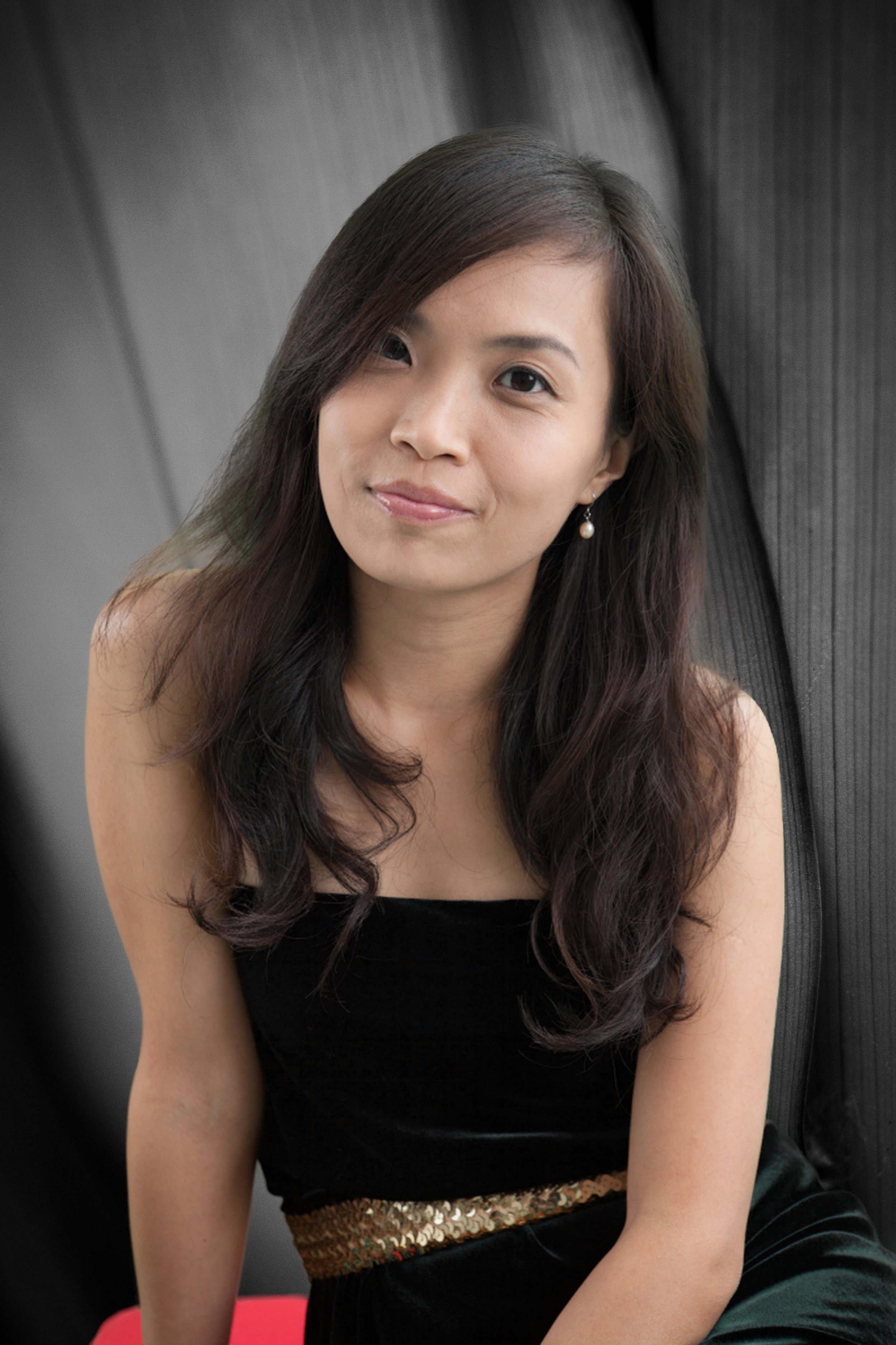A young woman with long wavy dark hair, wearing a black strapless dress with a gold sequin belt, sitting on a red surface against a dark grey textured background.