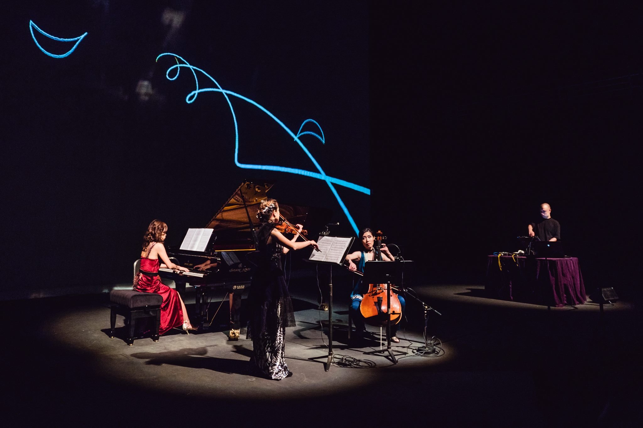 A group of musicians performing on stage with a dark backdrop. The group includes a pianist, a violinist, a cellist, and a person operating electronic equipment. There is a blue light projection in the shape of a whimsical, abstract design on the backdrop.