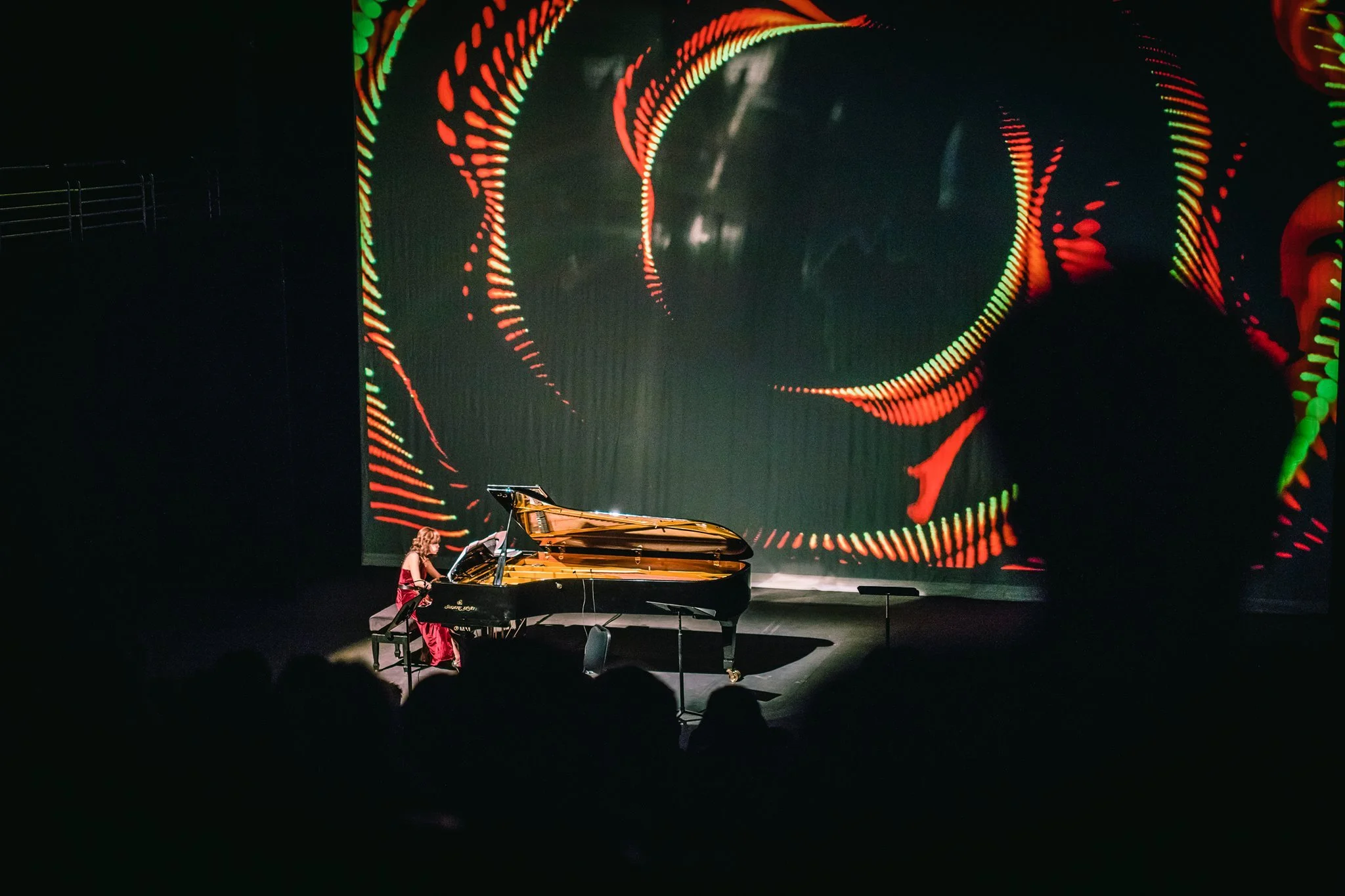 A young girl in a red dress playing a grand piano on a stage with a large digital backdrop displaying swirling red, green, and yellow abstract patterns.
