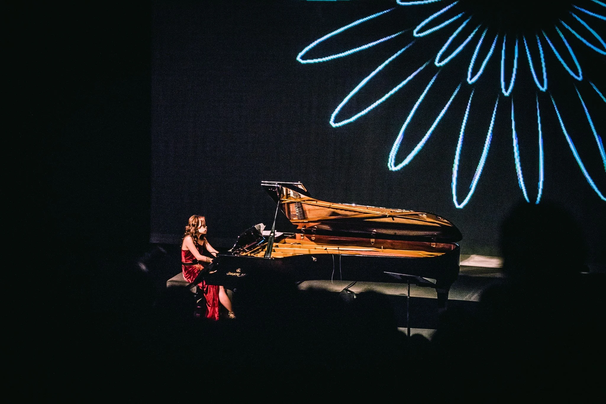 A young girl in a red dress playing a grand piano on a stage with a dark background. There is a large blue neon flower design projected on the wall behind her.