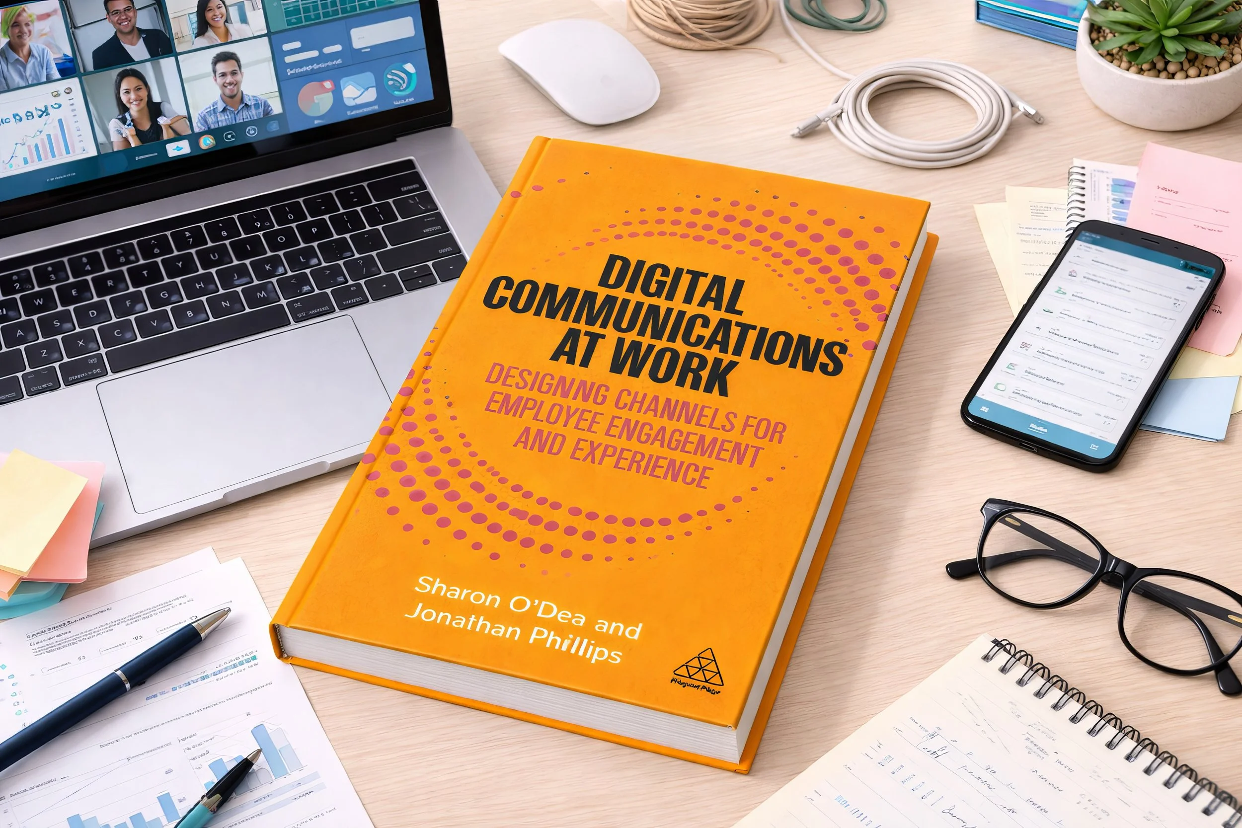 A workspace with a laptop, smartphone, glasses, a book titled 'Digital Communications at Work,' documents, a pen, sticky notes, and a potted plant on a light wooden desk.
