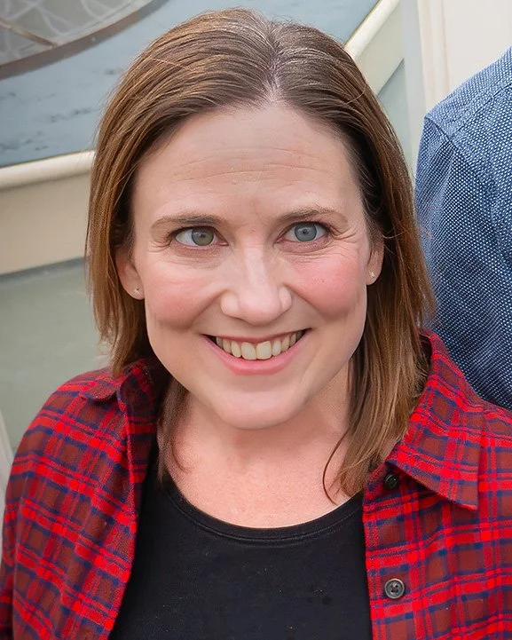 A woman with shoulder-length brown hair, blue eyes, and fair skin, smiling while wearing a red and black plaid shirt over a black top.