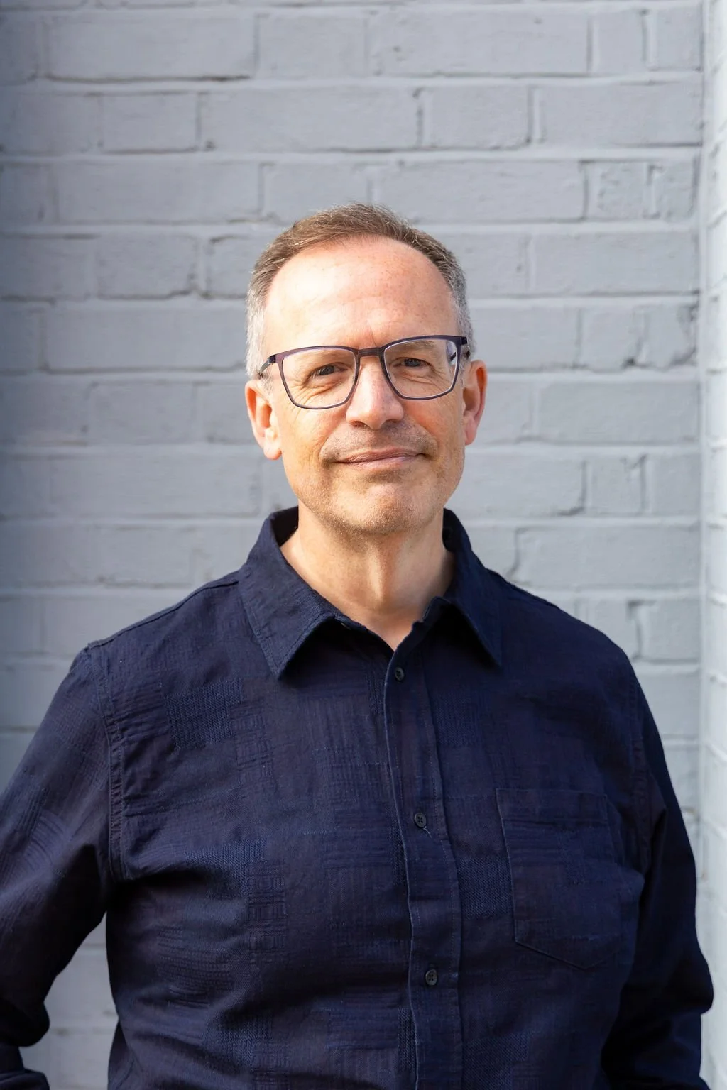 Jonathan Phillips, co-author of Digital Communications at Work, wearing a dark blue collared shirt standing in front of a light gray brick wall.