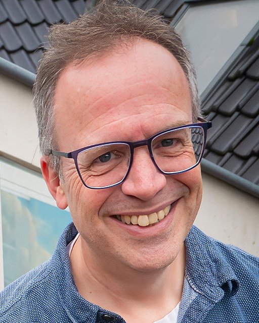 Smiling man with glasses outdoors, with buildings and a cloudy sky in the background.