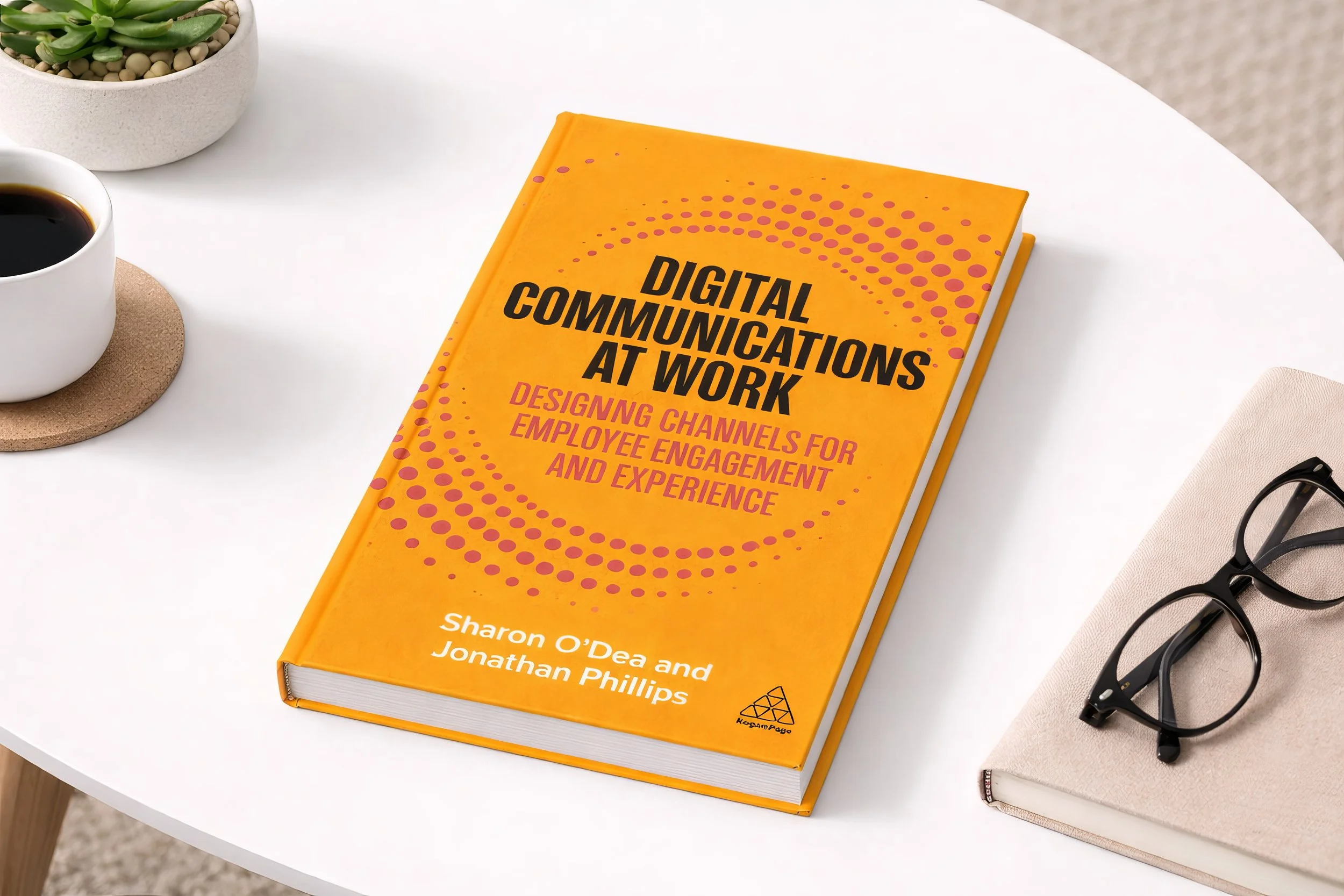 A book titled 'Digital Communications at Work' on a white table, alongside a cup of black coffee, a pair of black glasses on a closed beige notebook, and a small potted plant.