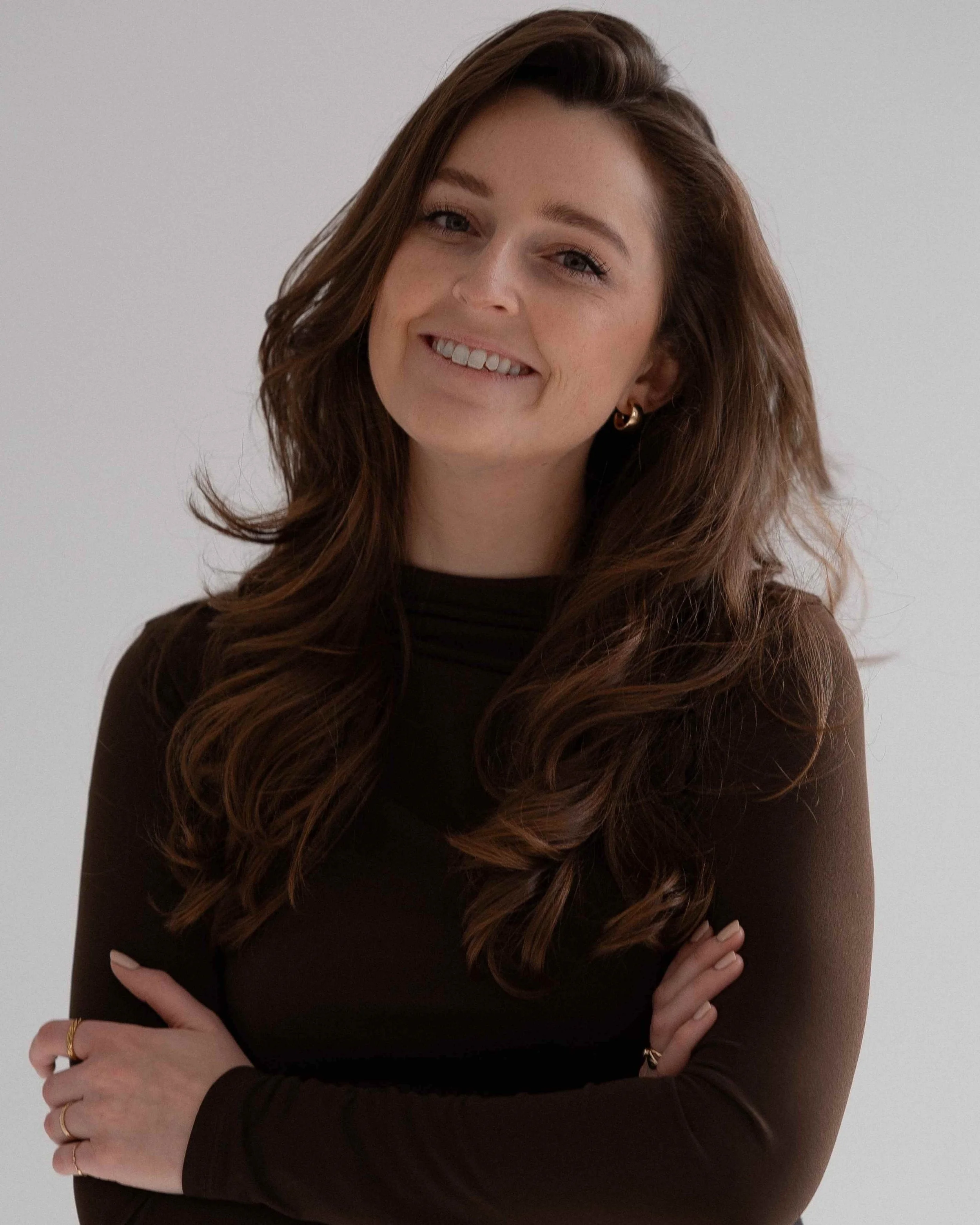 Anna-Sophie M. steht mit langen, lockigen braunen Haaren, schwarzem Kleid, Goldringen und Ohrringen vor hellem Hintergrund. Sie hat die arme leicht vor der Brust verschränkt.