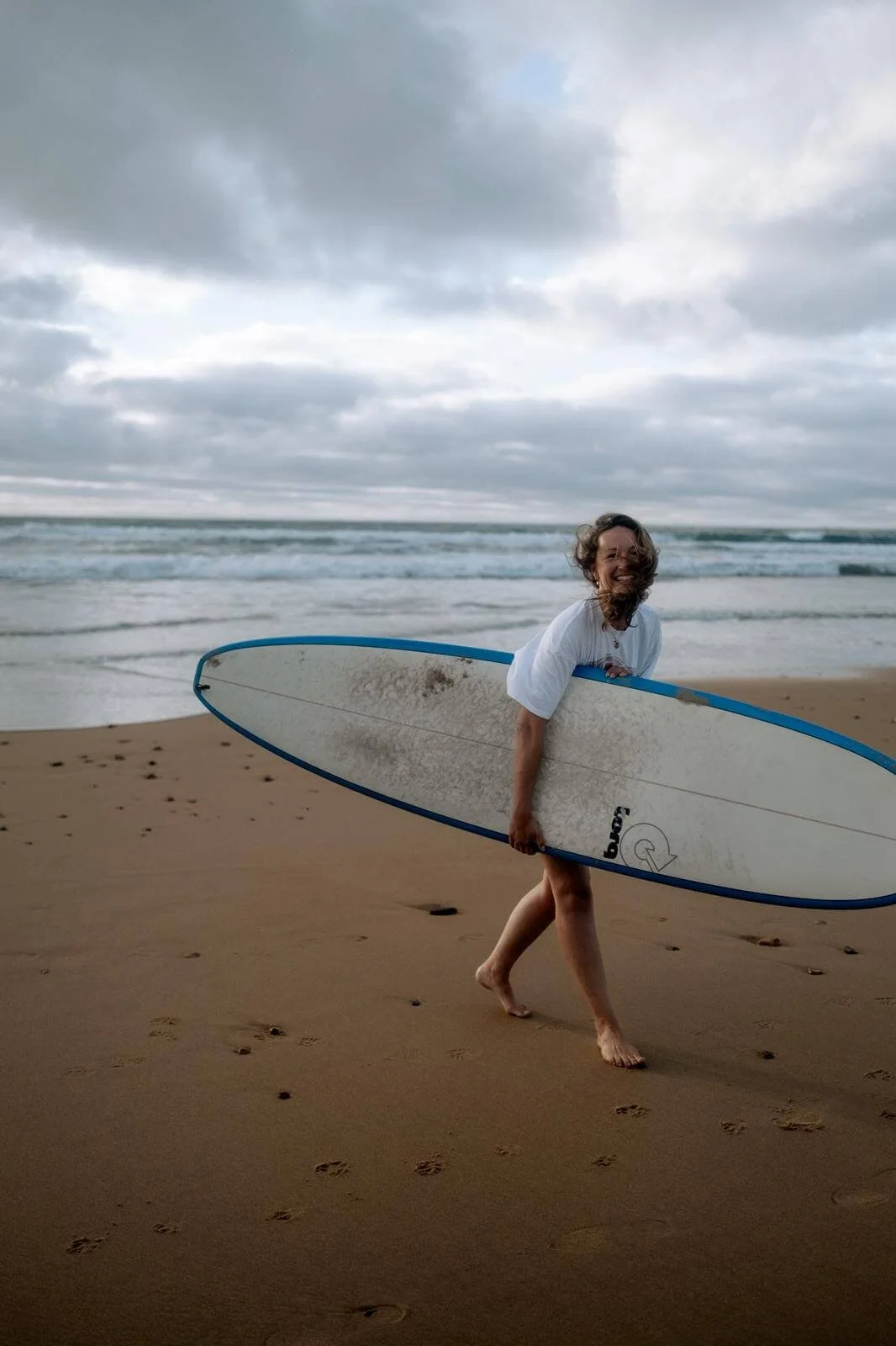 Jenna mit Surfbrett unter dem Arm am Strand bei bewölktem Himmel, lachend, trägt weiße Kleidung, barfuß auf Sand, Meereswellen im Hintergrund.