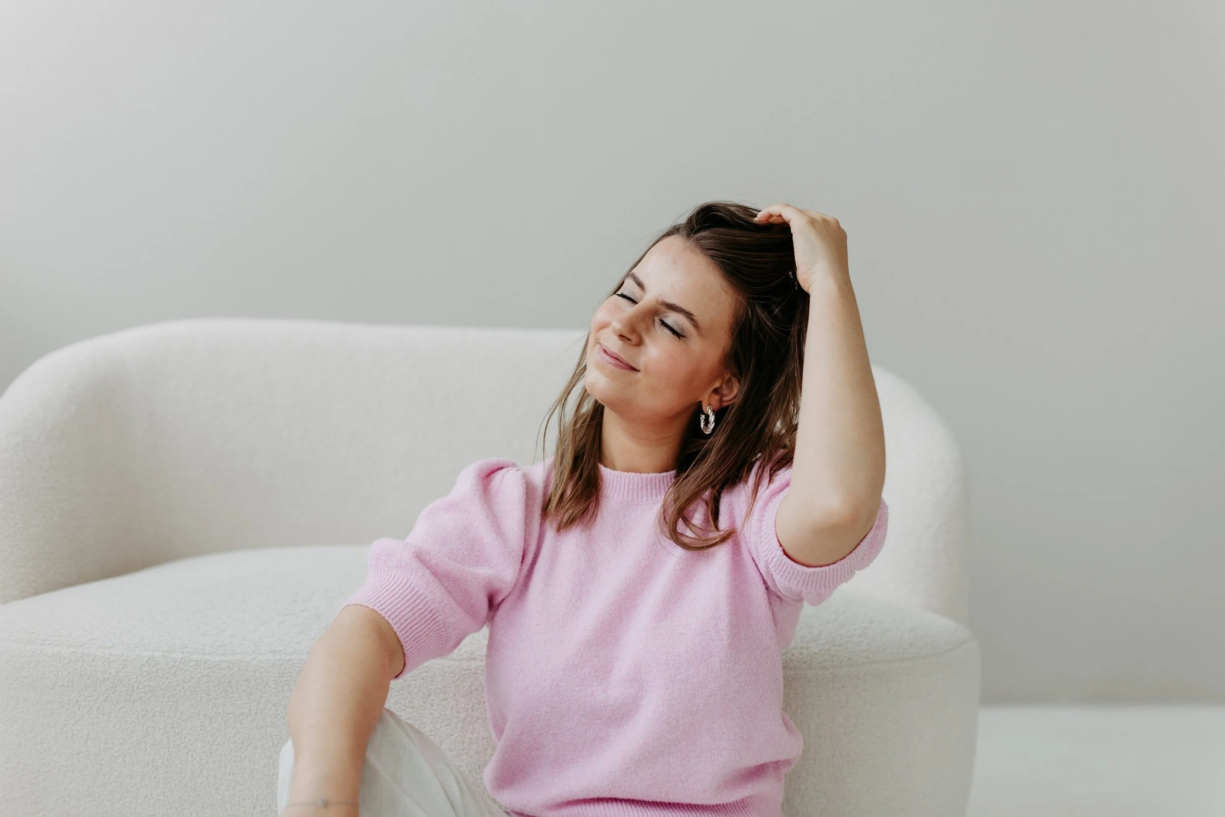Kristin sitzt in einem rosa T-shirt vor einem weißen Sofa das vor einem hellgrauen Hintergrund steht, hat die Augen geschlossen und lächelt, während sie den Kopf mit der Hand berührt.
