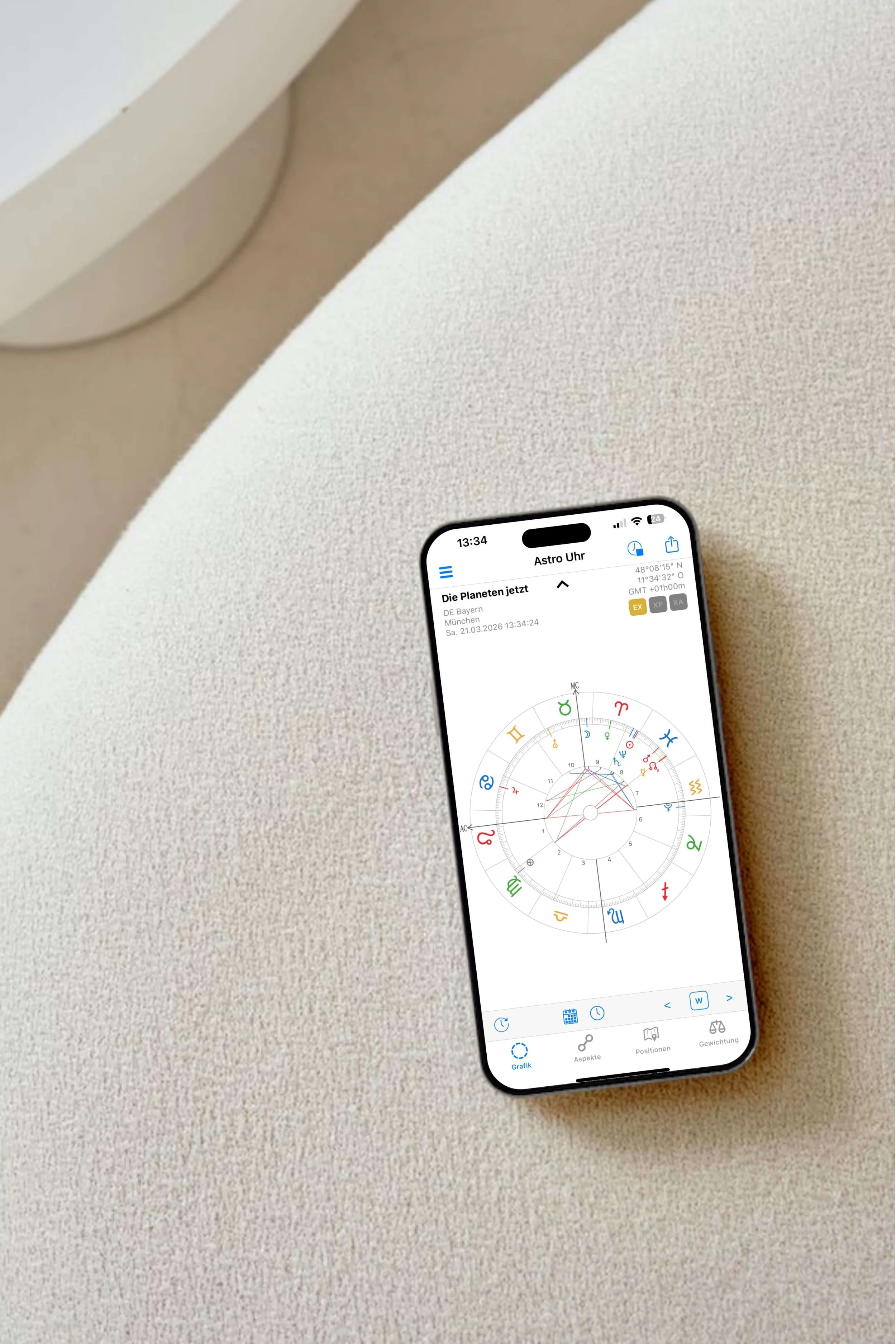 Ein Smartphone mit einer Astro-App liegt auf einem hellen, strukturierten Sofa. Auf dem Bildschirm ist ein Geburtshoroskop sichtbar.