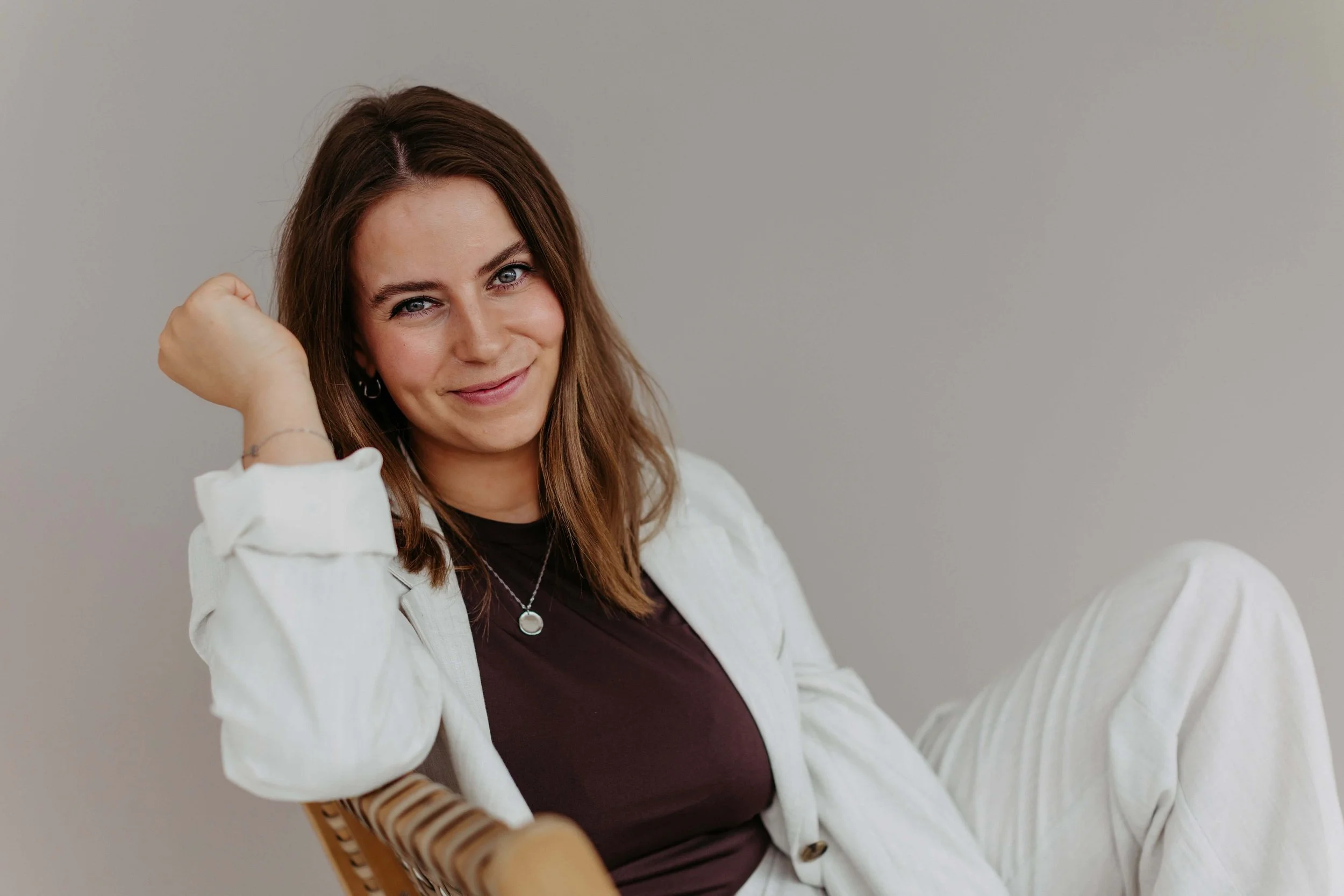 Kristin sitzt entspannt in einem hellen Outfit auf einem Stuhl und lächelt freundlich in die Kamera.