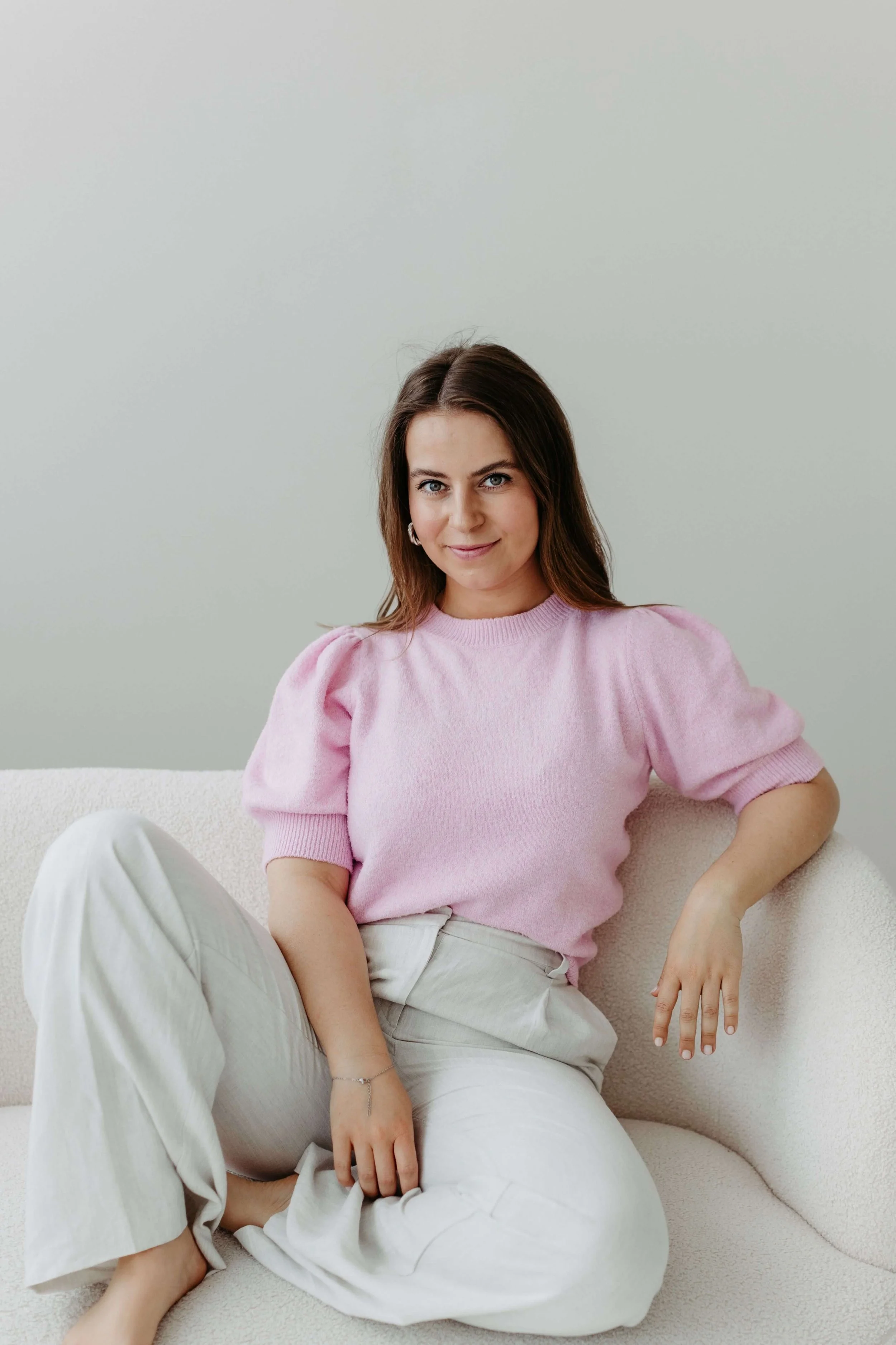 Kristin sitzt entspannt auf einem weißen Sofa, trägt einen pinken Pullover und weiße Hosen, hat braune Haare und blickt in die Kamera.