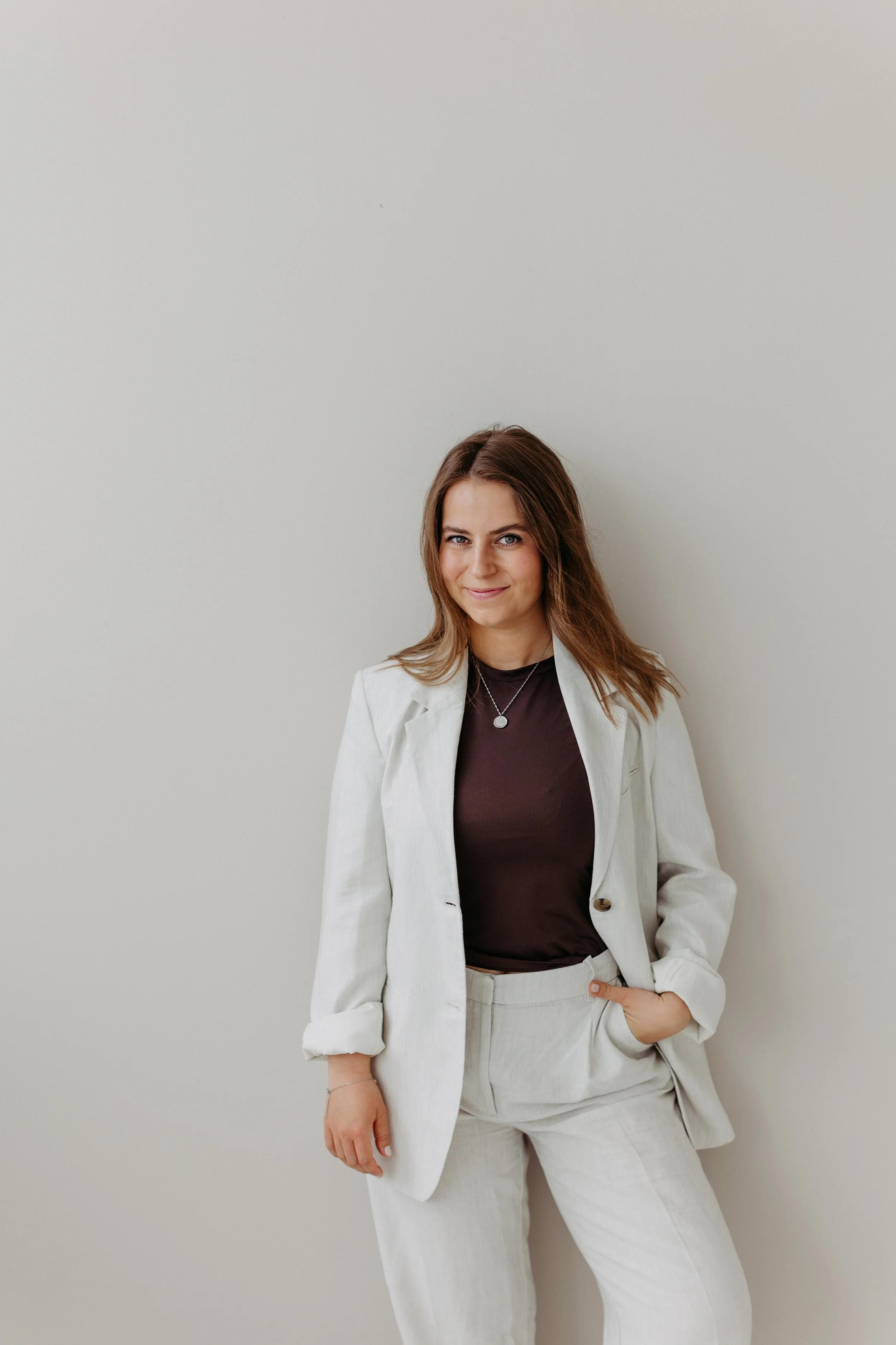 Junge Frau in weißem Anzug und braunem Shirt, die vor einer weißen Wand steht.