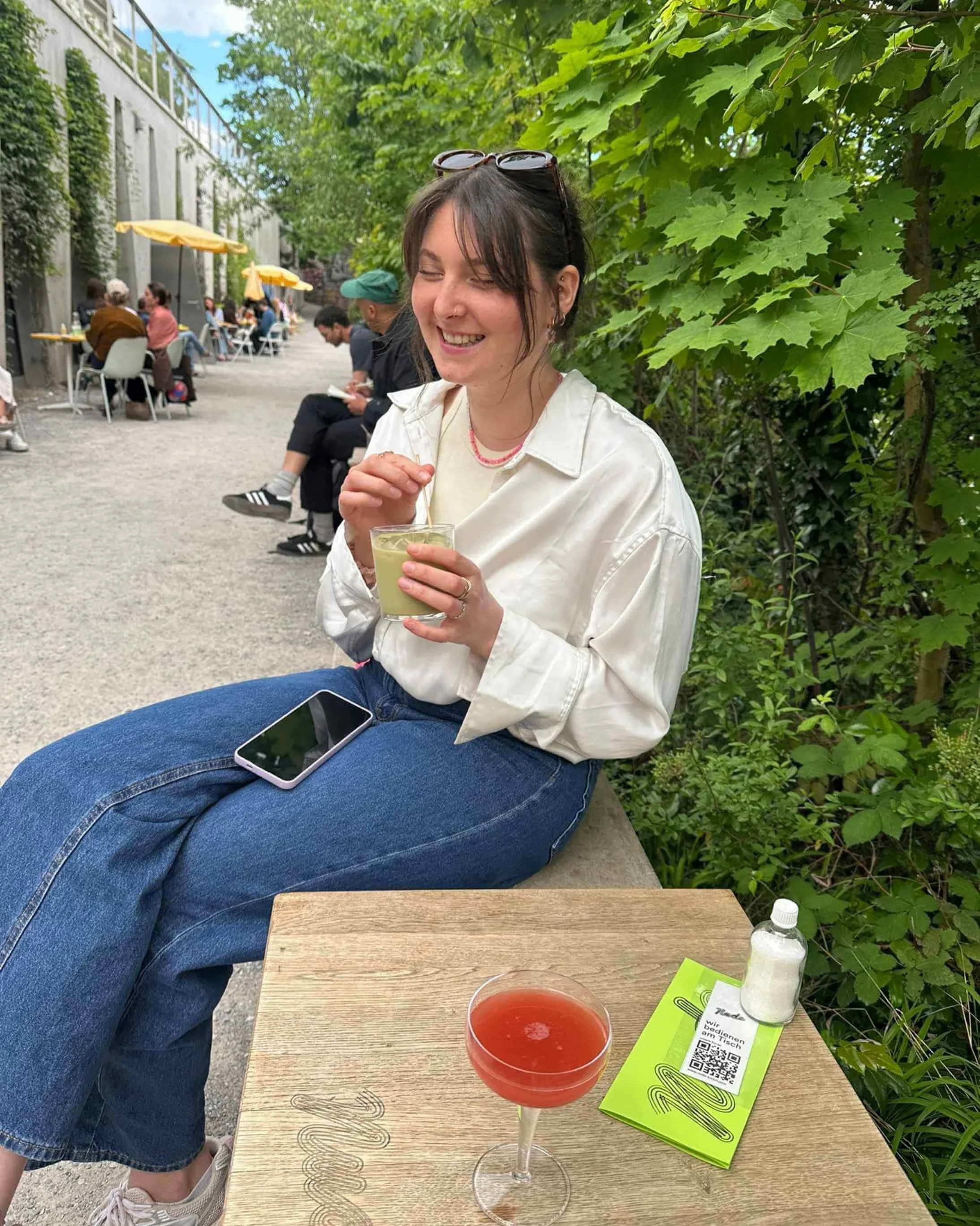 Anni N. mit braunen Haaren, Sonnenbrille auf Kopf, im weißen Hemd, sitzt im Park mit Getränk, Smartphone auf dem Schoß, neben einem Tisch mit einem roten Getränk, einer Flasche und einem Flyer.