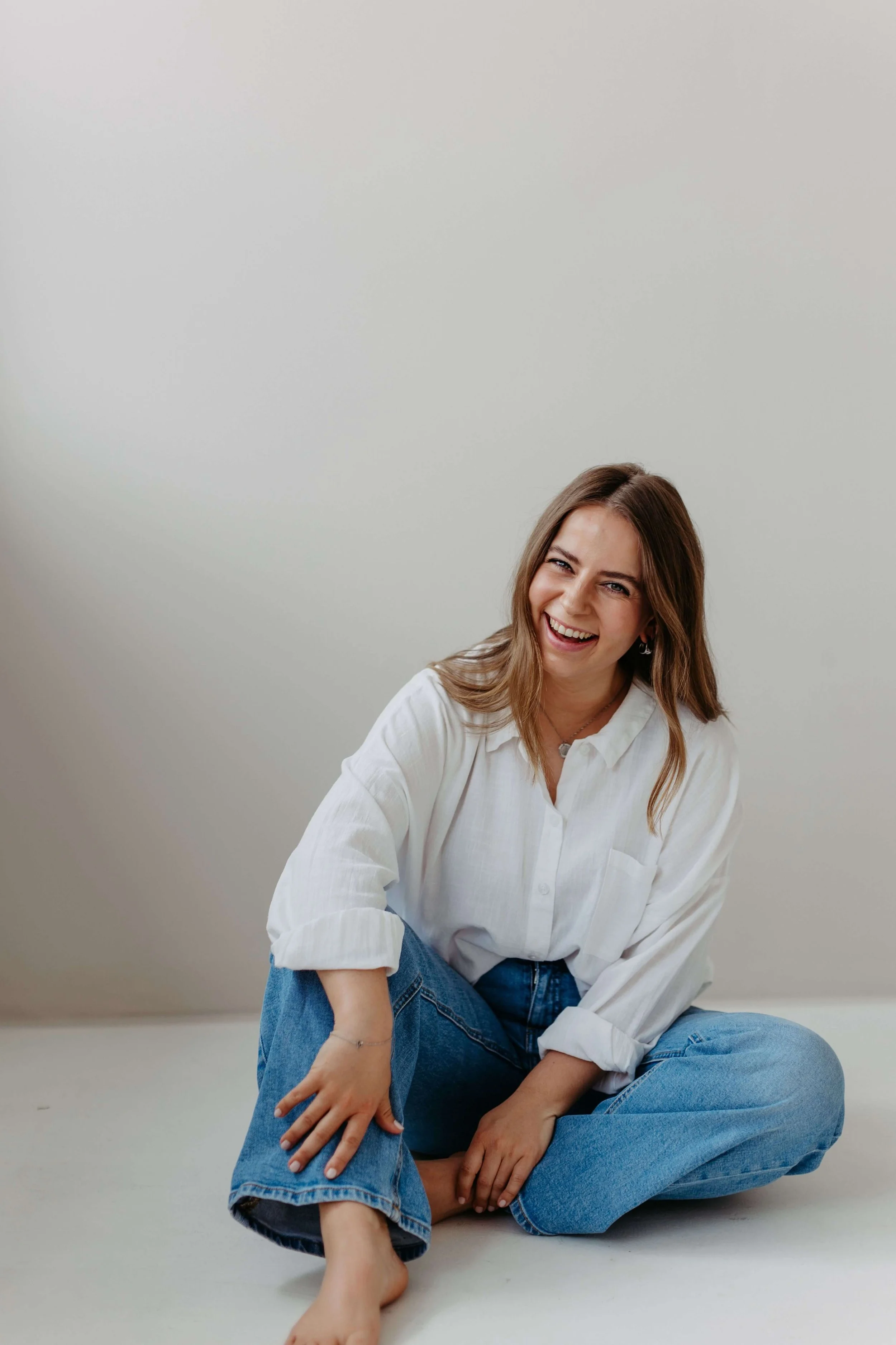 Kristin sitzt lachend mit dunkelblonden Haaren, in weißem Hemd und blauen Jeans, barfuß auf dem Boden vor einer weißen Wand.
