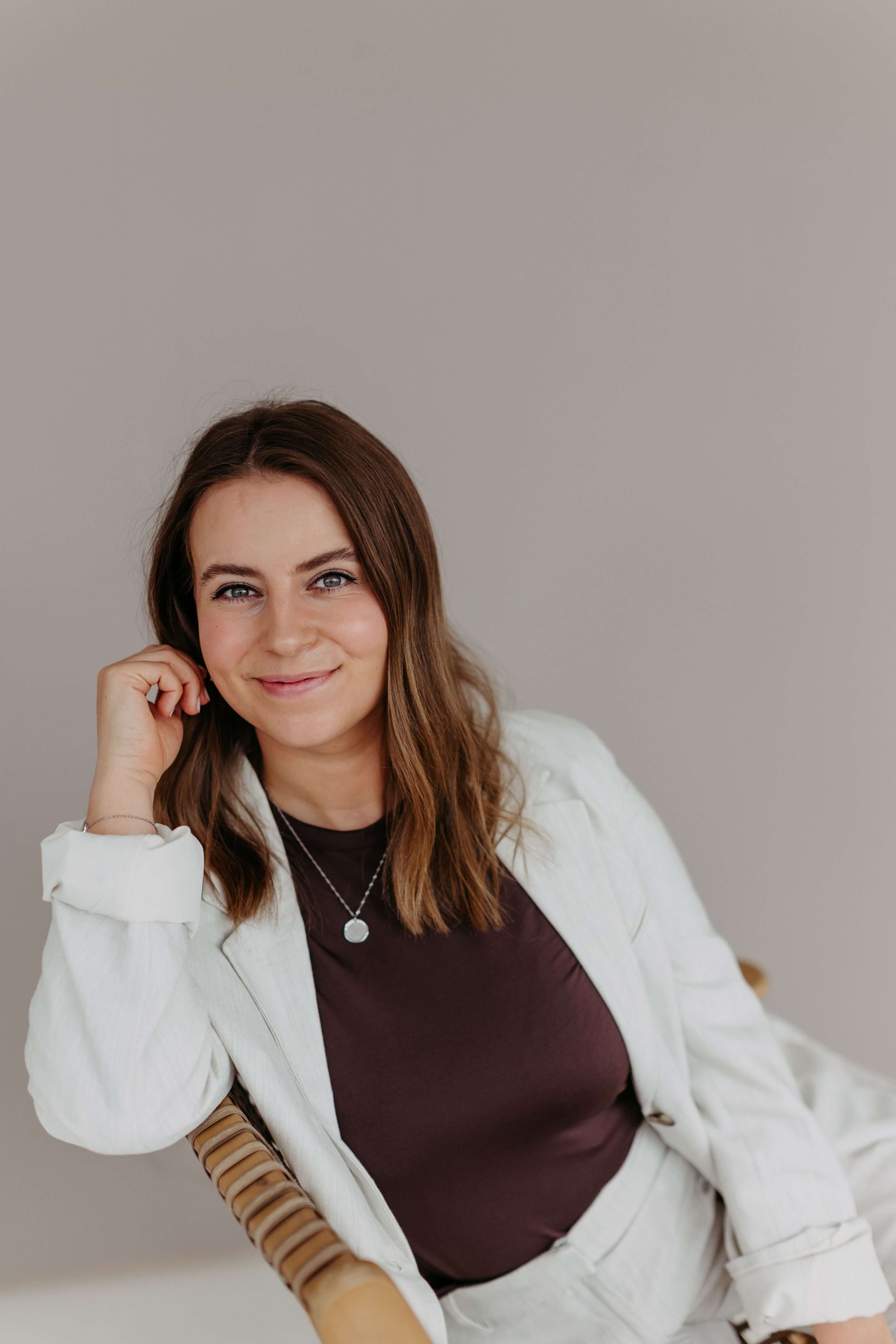Kristin mit dunkelblonden, gewellten Haaren sitzt auf einem Stuhl, trägt ein helles Jackett, ein dunkelbraunes T-Shirt und eine Halskette, lächelt in die Kamera und hat die Hand an ihr Gesicht gelegt.