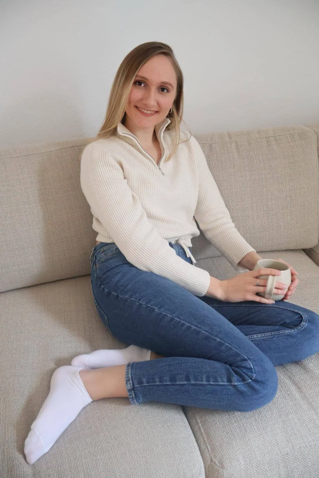 Verena S. sitzt auf einem Sofa, trägt weiße Pullover und blaue Jeans, hält eine Tasse Kaffee und lächelt in die Kamera.