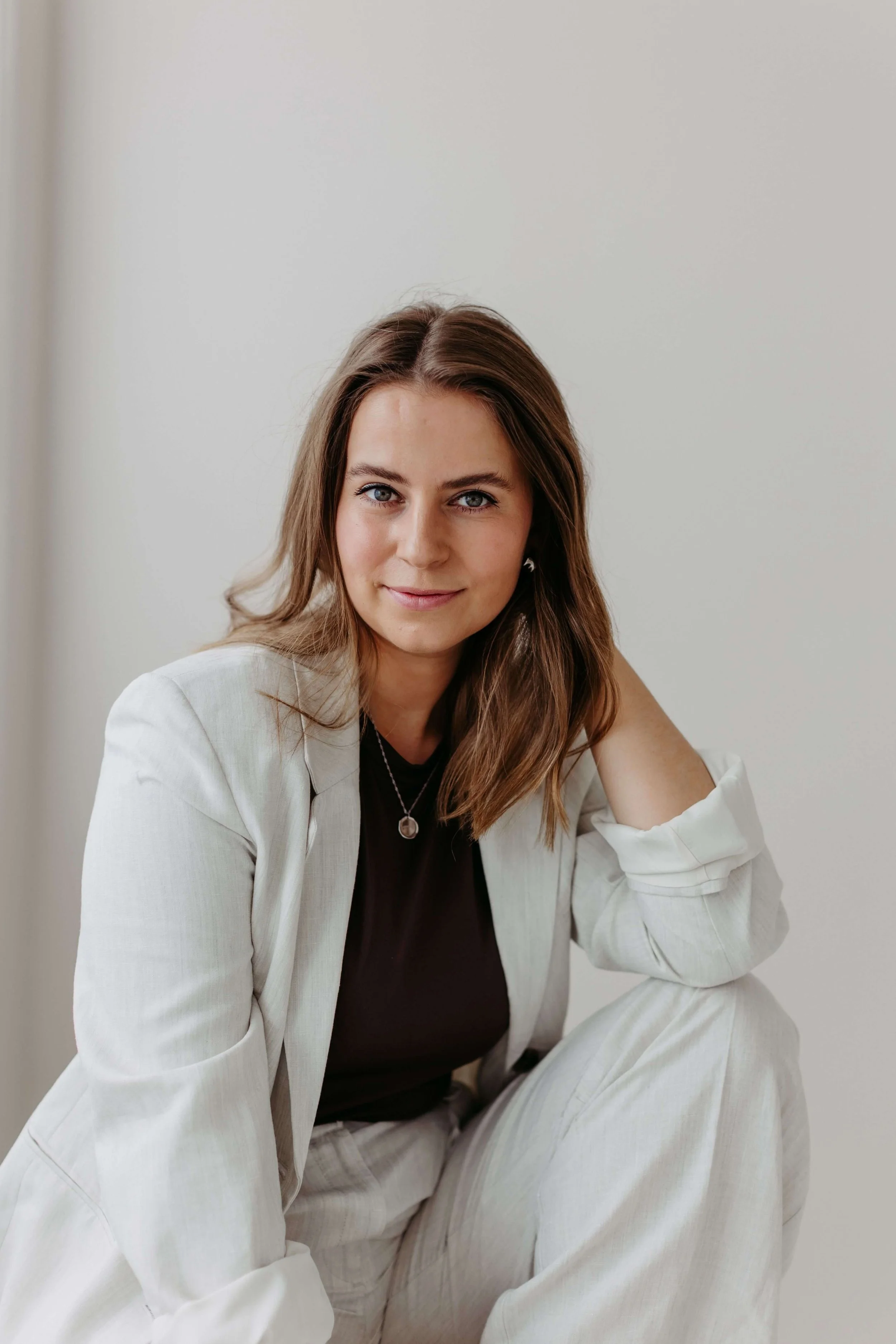 Kristin, mit dunkelblonden Haaren, trägt ein dunkelbraunes shirt und einen hellen Blazer mit passender Hose und sitzt vor einer weißen Wand. Die eine Hand auf dem Knie abgestützt und schaut lächelnd in die Kamera.