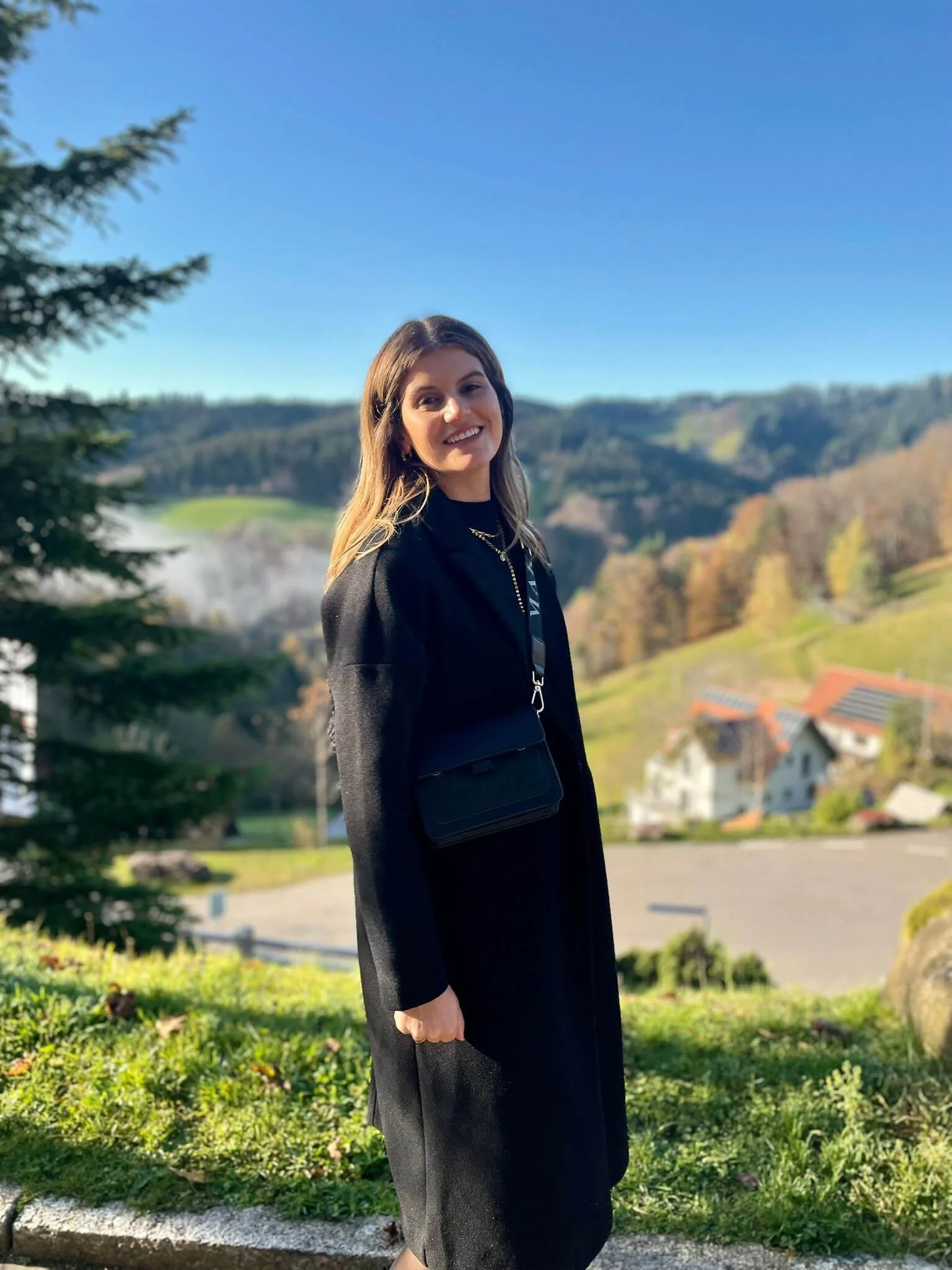 Eine junge Frau steht im Freien vor einer malerischen Landschaft mit Hügeln, Bäumen und Häusern, das Wetter ist sonnig und klar.