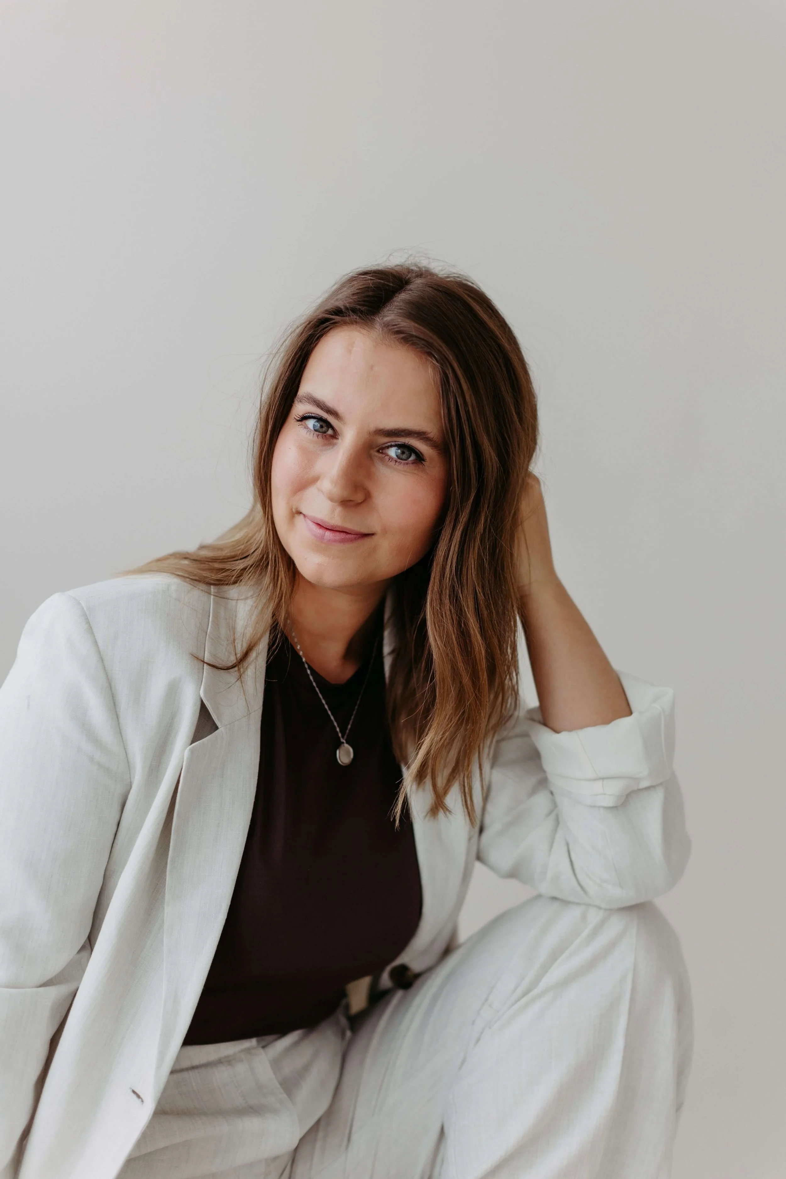 Kristin schaut mit freundlichem Lächeln, in die Kamera. Sie trägt einen hellen Blazer, einen dunkelbraunes Oberteil und eine Halskette, sitzt vor einer neutralen Wand, lächelt leicht und schaut freundlich in die Kamera.