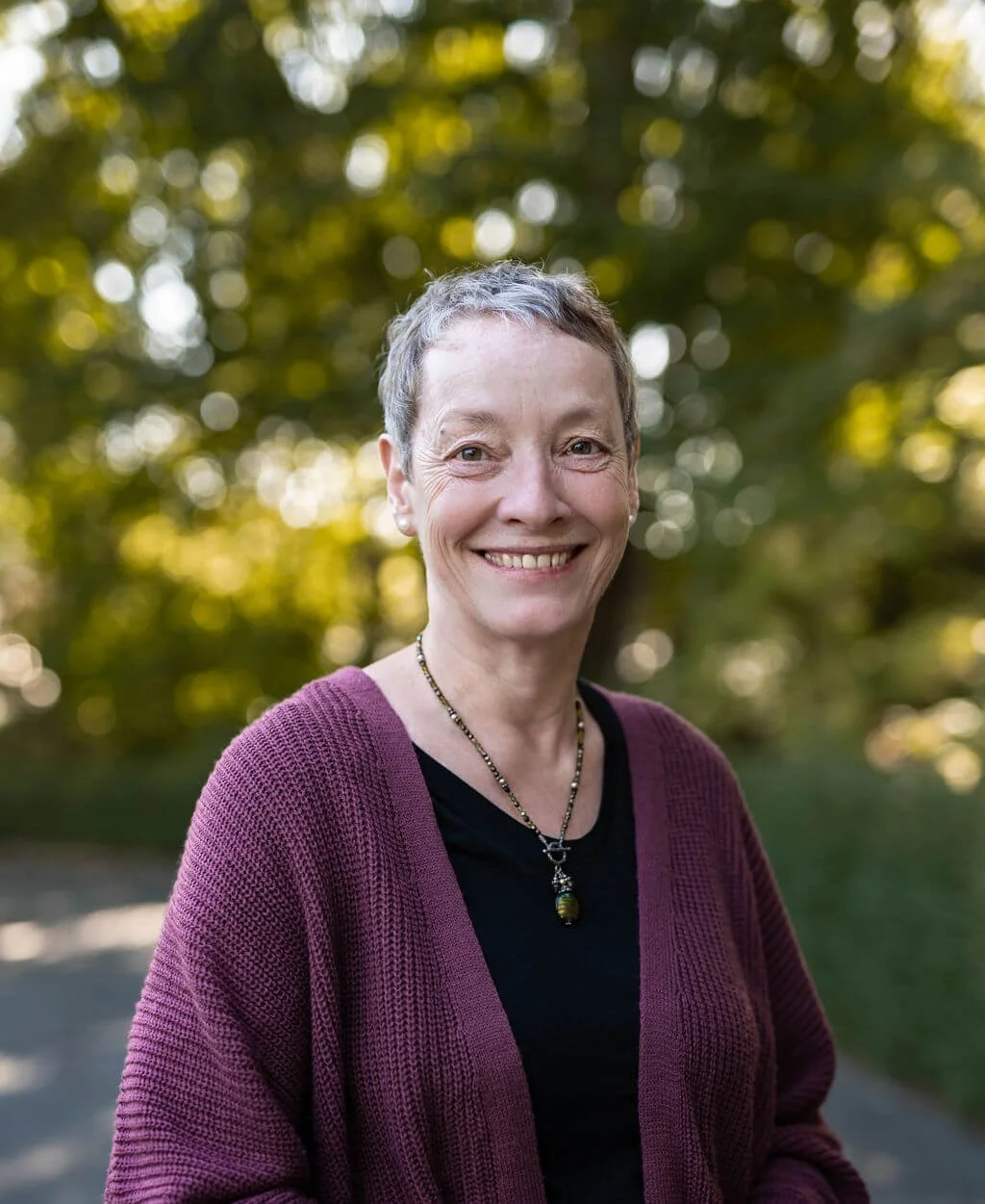 Marion M. lächelt in die Kamera, hat kurze graue Haaren, trägt eine schwarze Bluse, einen lila Cardigan und eine Halskette, steht im Freien mit einem Hintergrund aus grünem, sonnigem Laub.