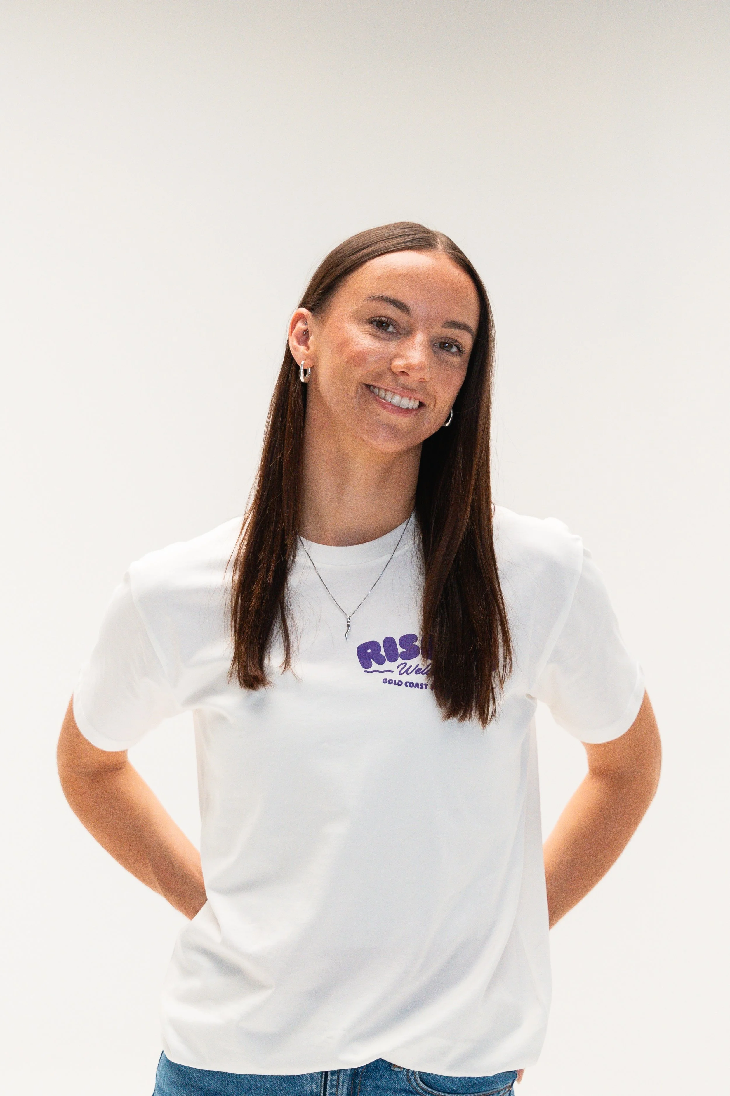 A young woman with brown hair wearing a white T-shirt and blue jeans standing against a plain white background.