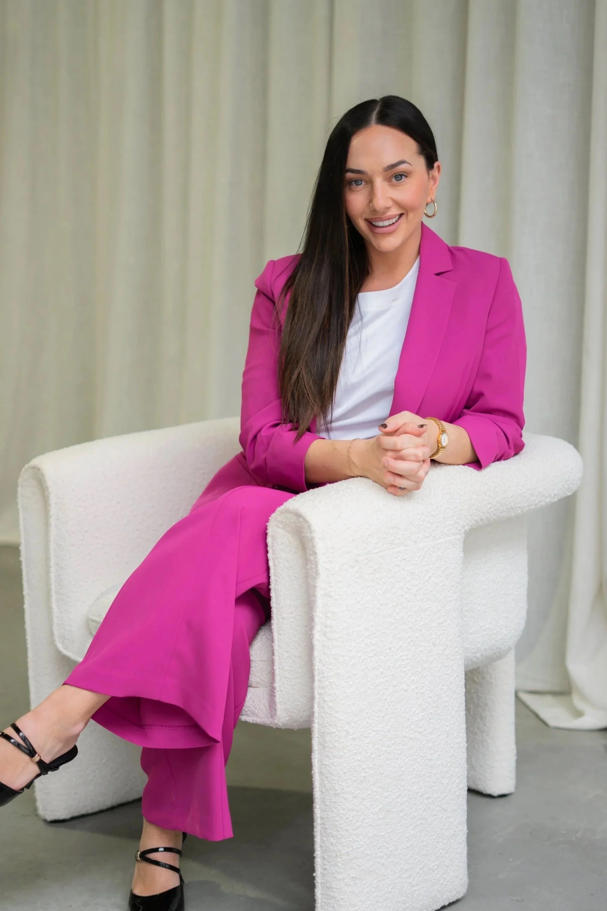 A woman with long dark hair, wearing a bright pink blazer and matching pants, sitting on a white textured chair with a modern design, smiling and looking at the camera.