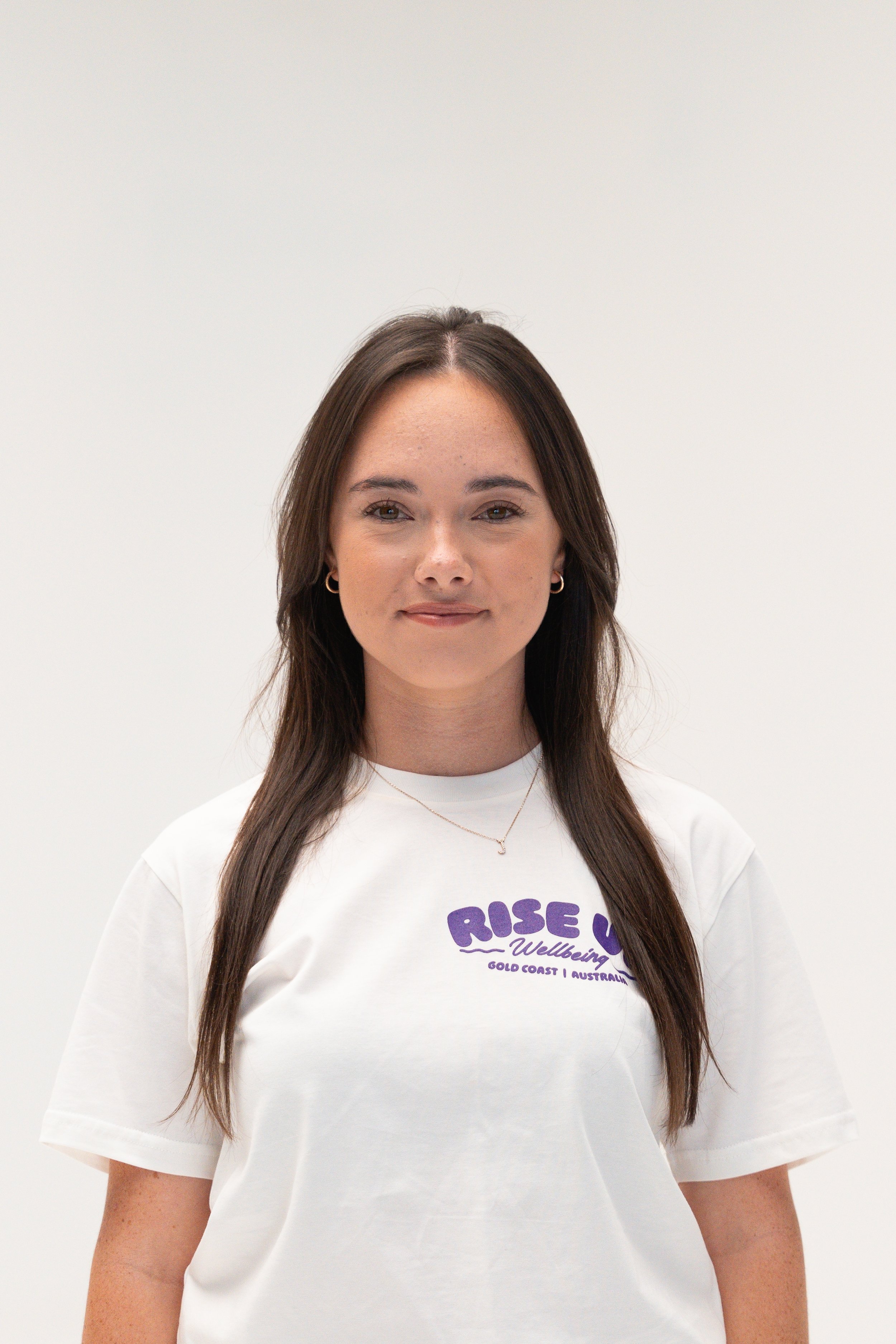 A young woman with long brown hair and hoop earrings, wearing a white t-shirt with purple text, standing against a plain white background.