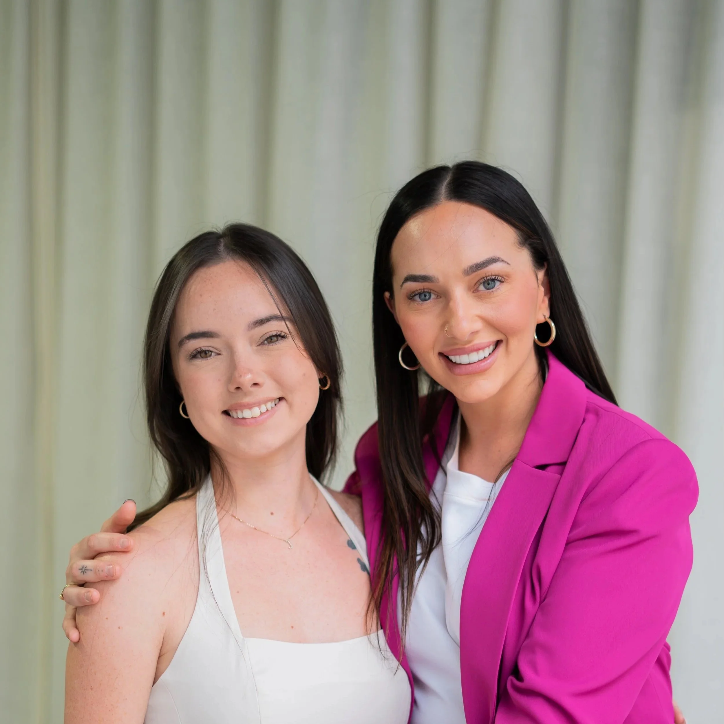 Two young women smiling, standing close together with one arm around each other's shoulders, against a light-colored curtain background.