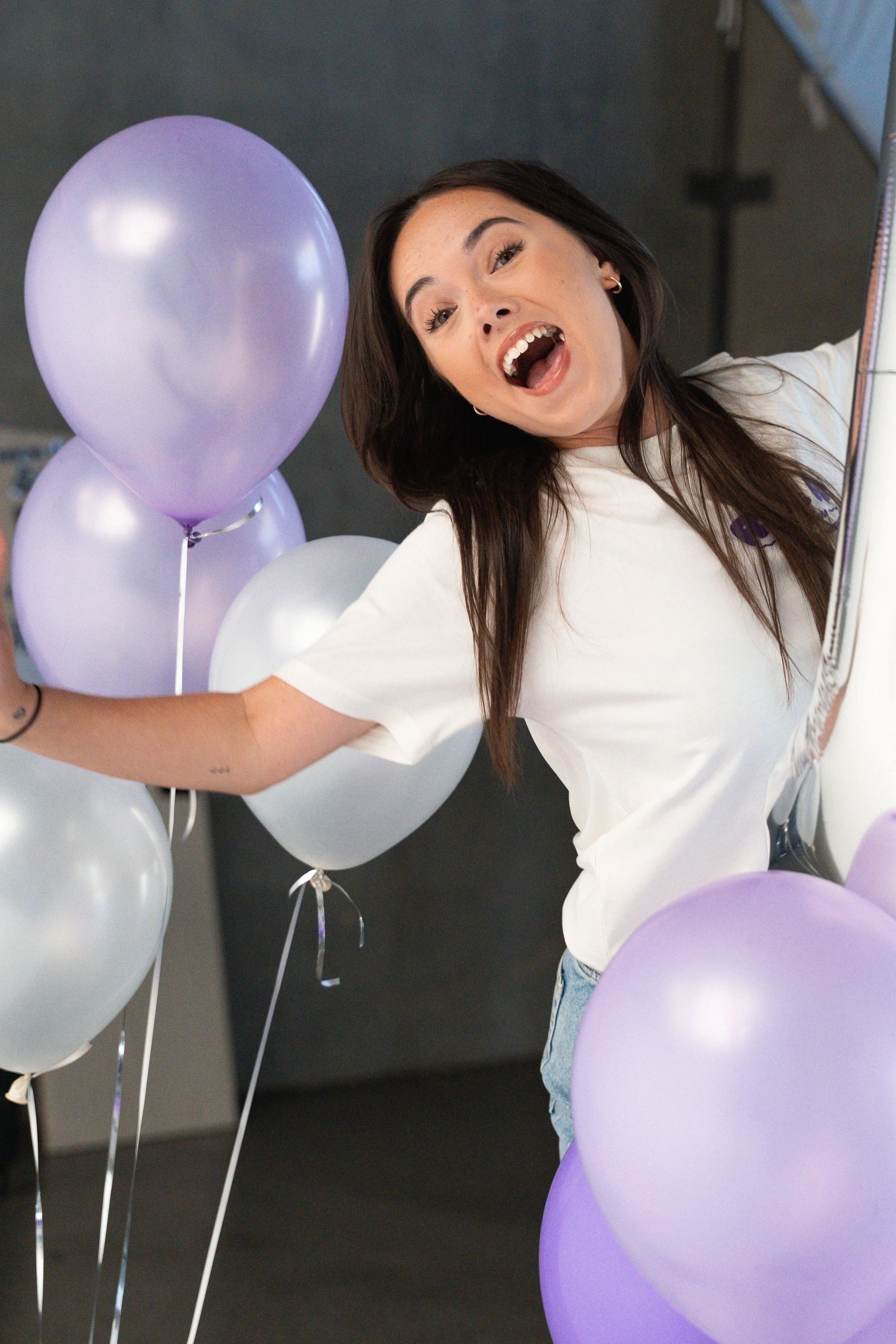 A woman with long dark hair smiling and reaching out among light purple and white balloons, with a gray background.