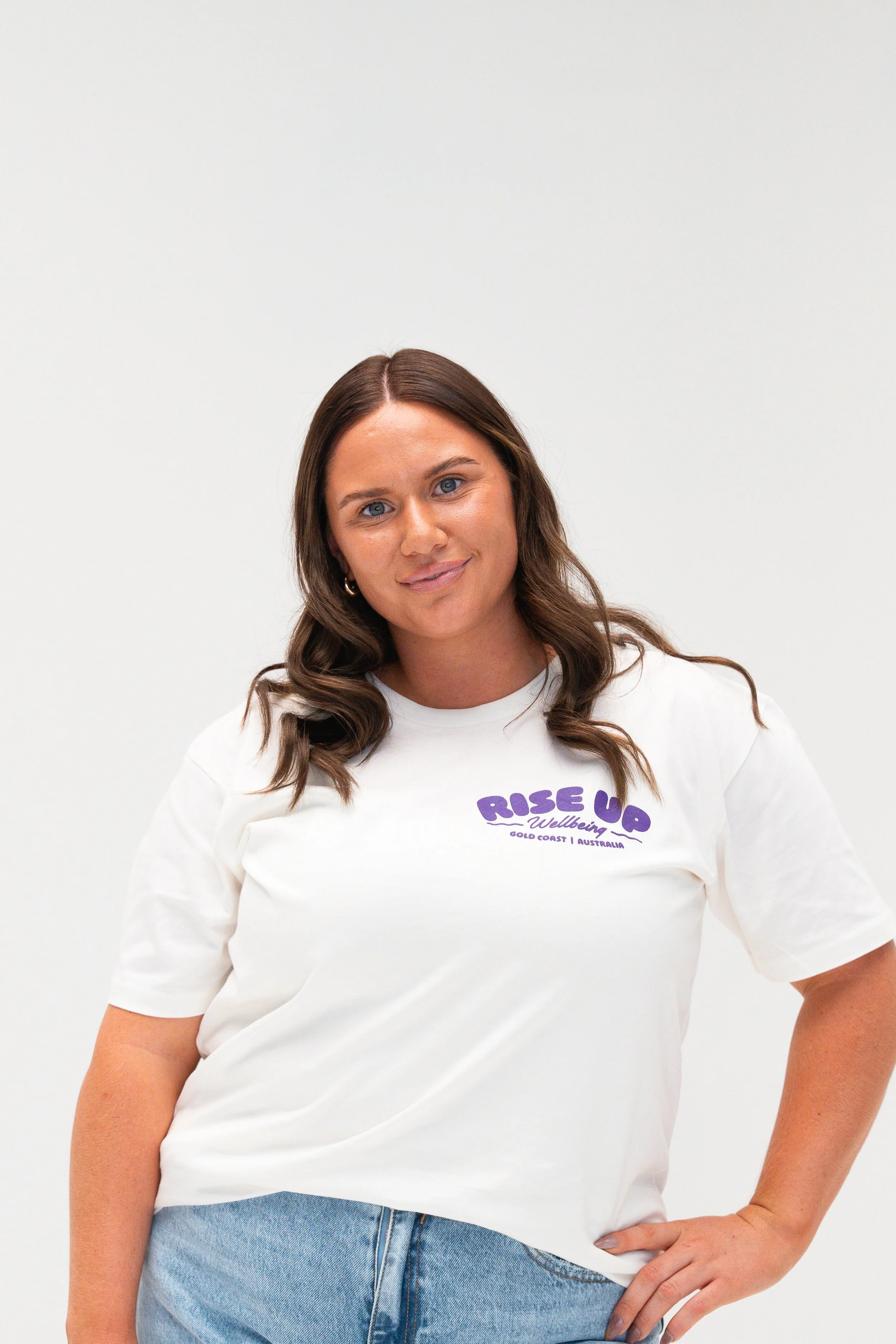 A woman with long brown hair and blue eyes wearing a white T-shirt with purple text and light blue jeans, standing against a plain white background.