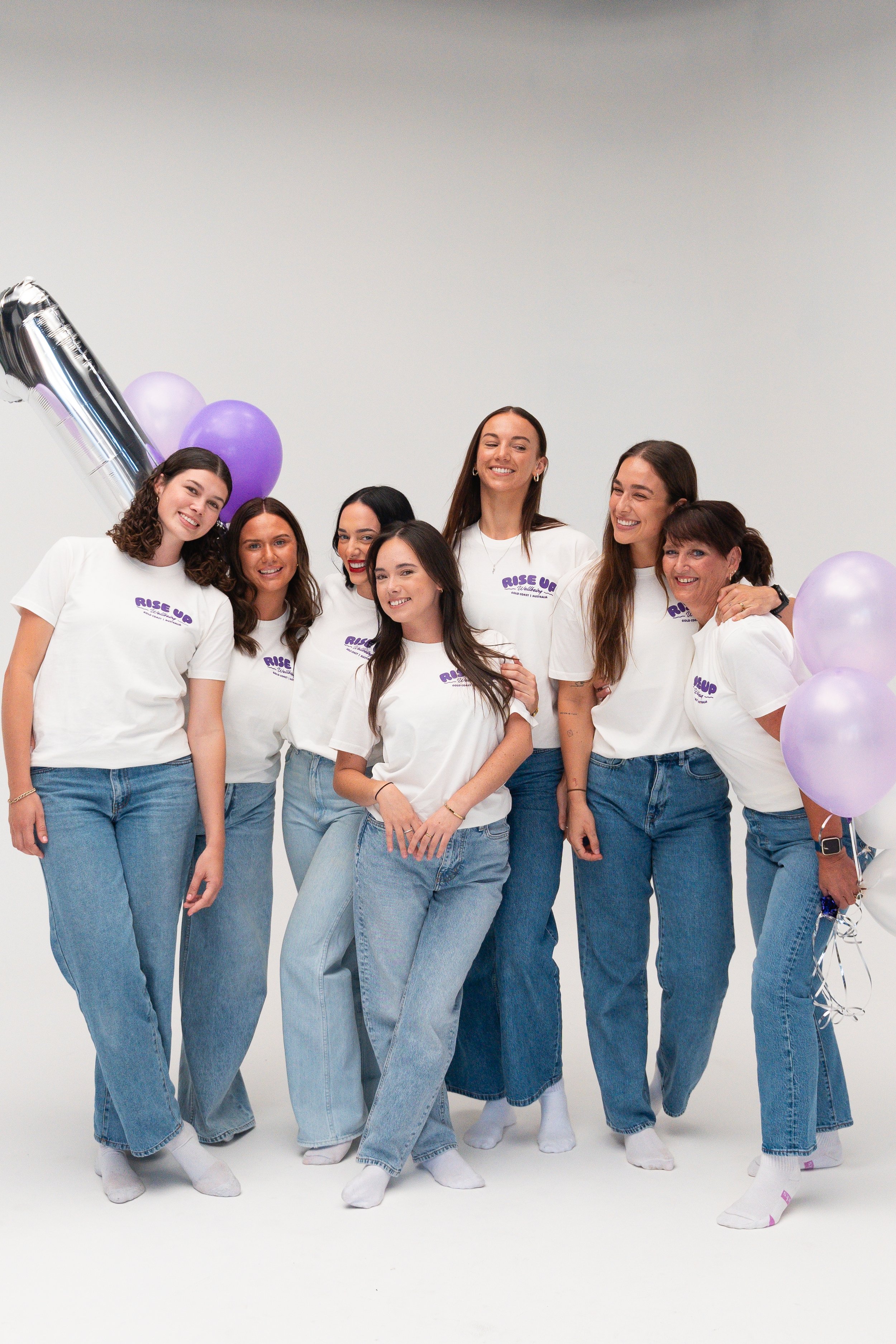 A group of seven women standing together in casual jeans and white T-shirts with "Rise Up" written on them, smiling at the camera, surrounded by purple and silver balloons, in a white studio setting.