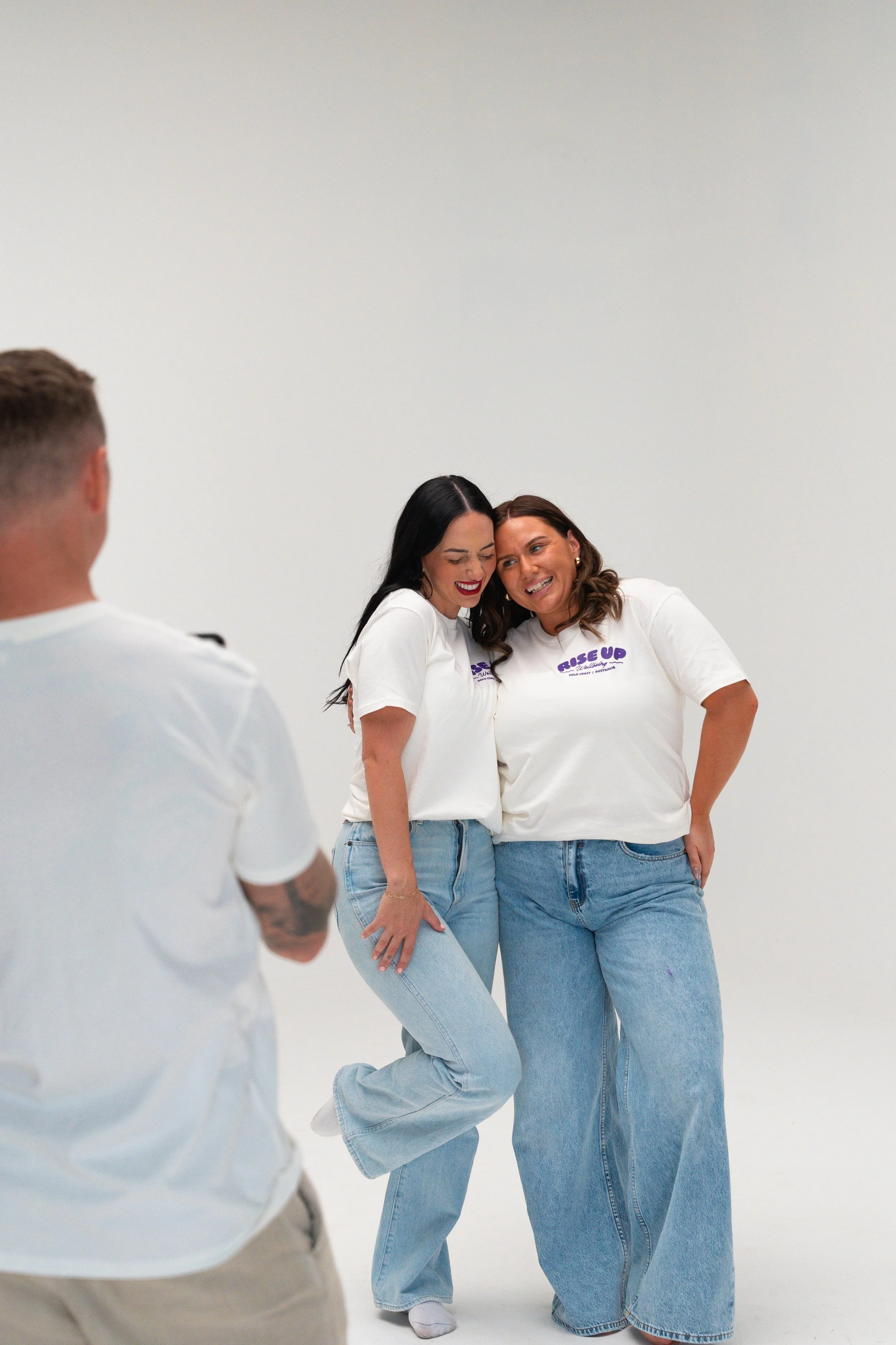 Two women are posing and laughing together in a white studio, wearing white t-shirts and blue jeans, with a man standing in the foreground, blurred, facing them.