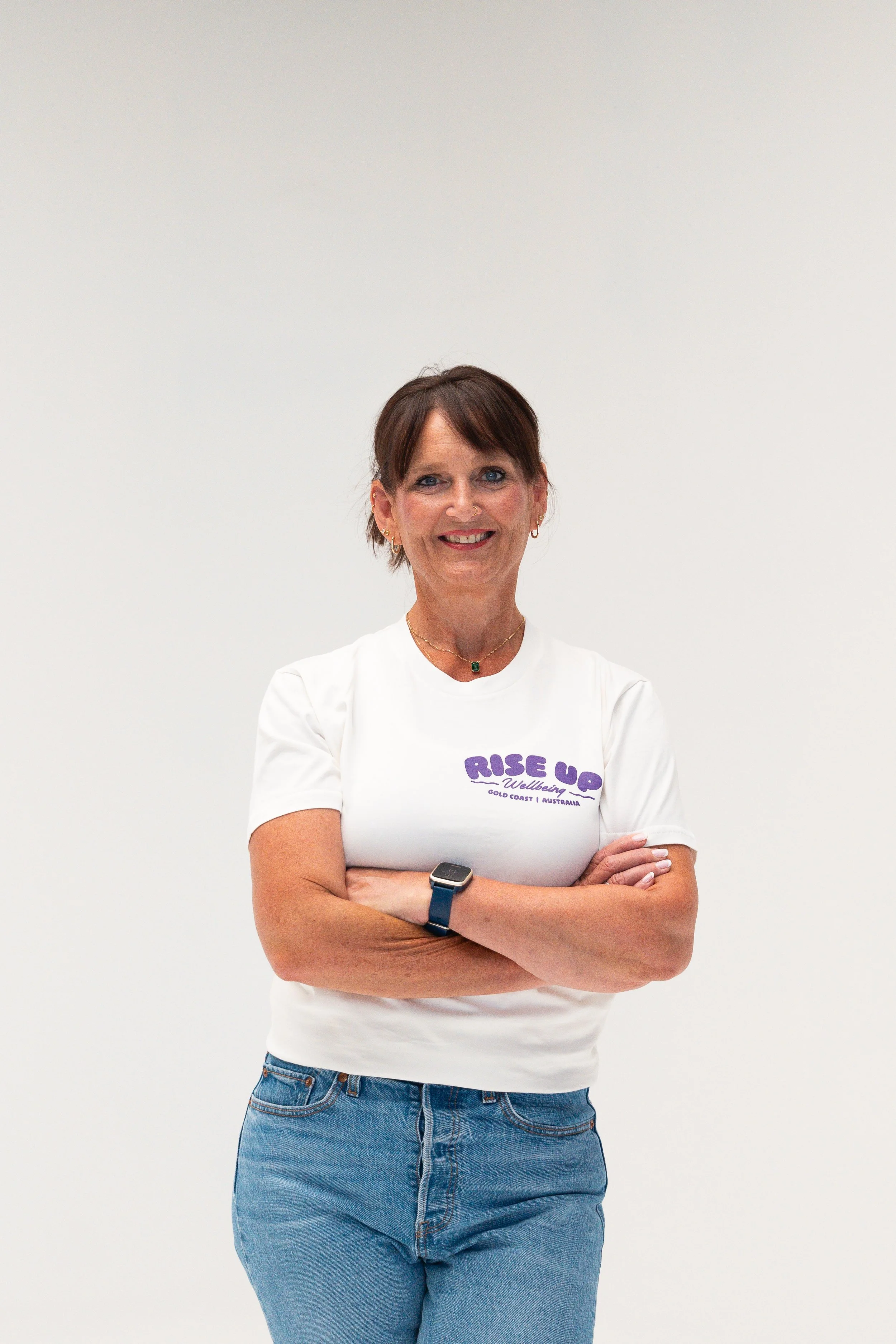 A woman with short brown hair, blue eyes, and earrings, smiling with arms crossed, wearing a white T-shirt with purple lettering and blue jeans, standing against a plain white background.
