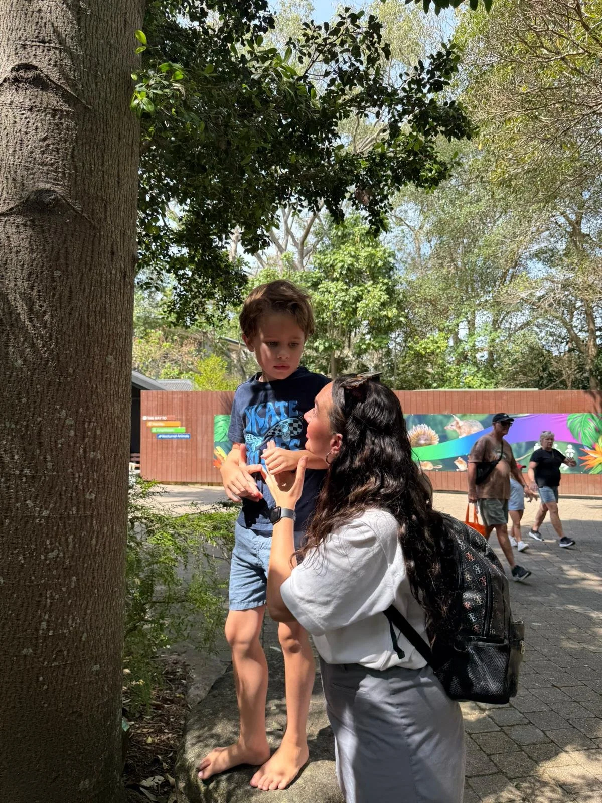 A woman with long dark curly hair and a backpack kneels on the ground, talking to a young boy standing barefoot on a rock next to a large tree, with other people walking in the background.