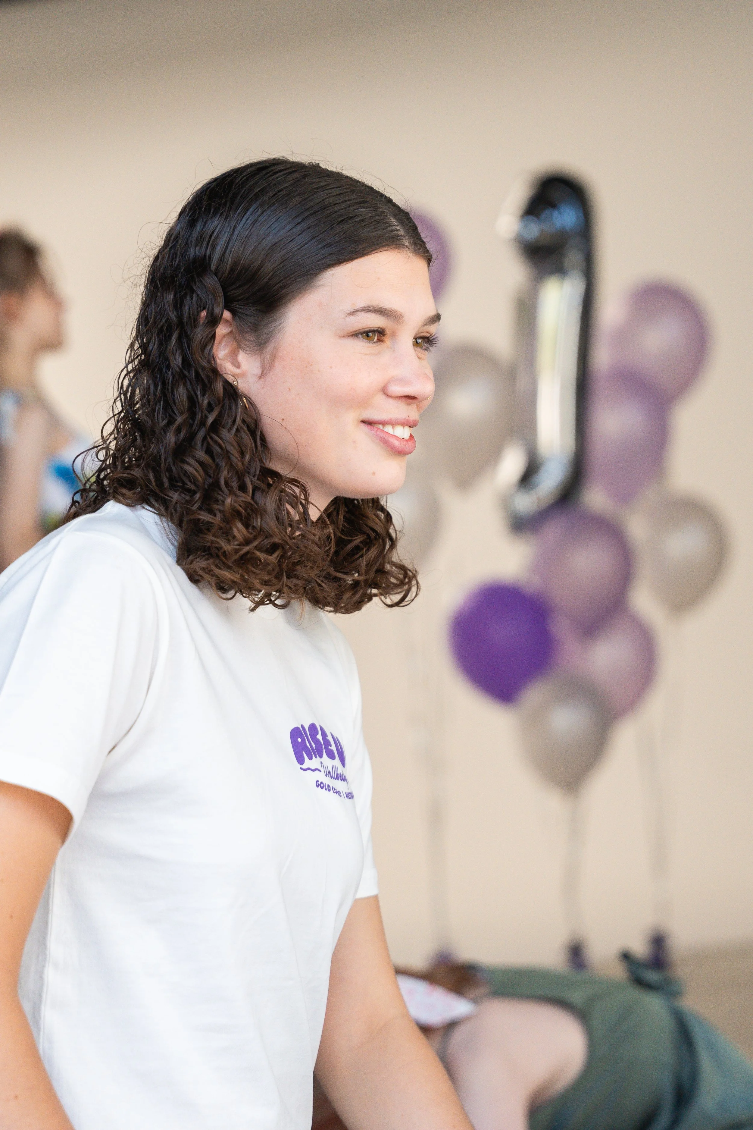 Young woman with brown, curly hair smiling in a room decorated with purple balloons.