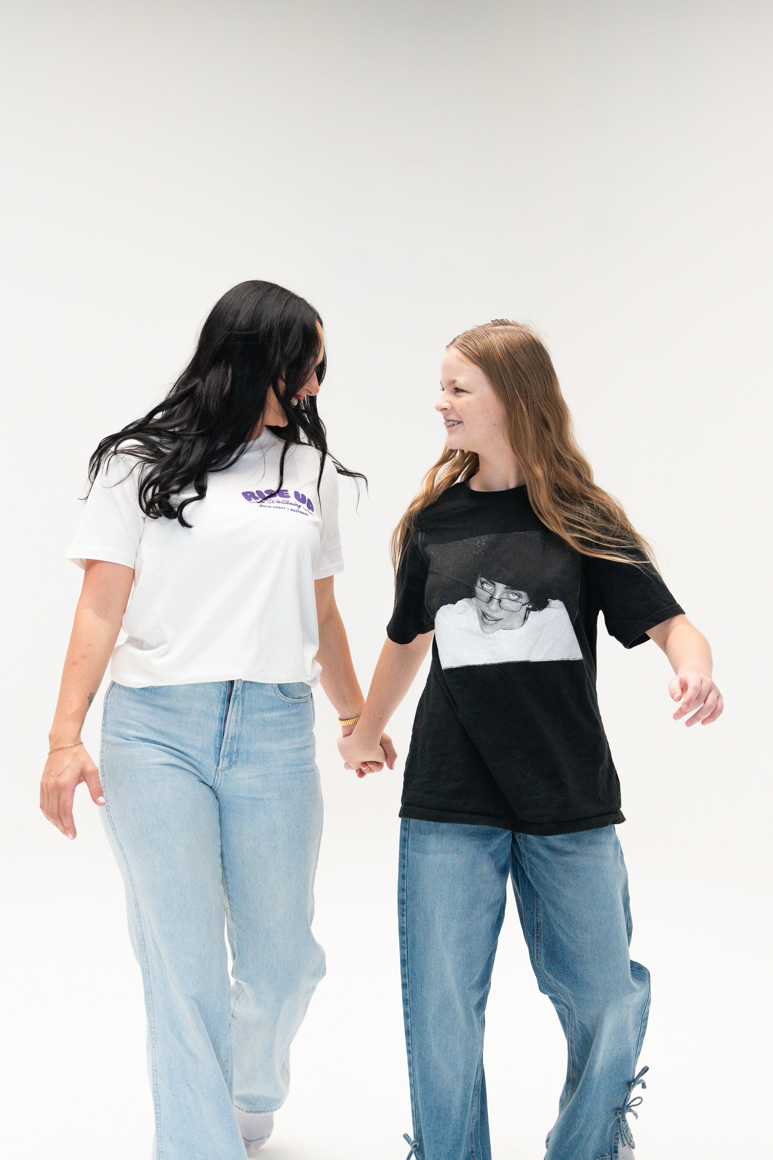 Two women holding hands and smiling at each other while walking together, dressed in casual clothing, against a plain white background.