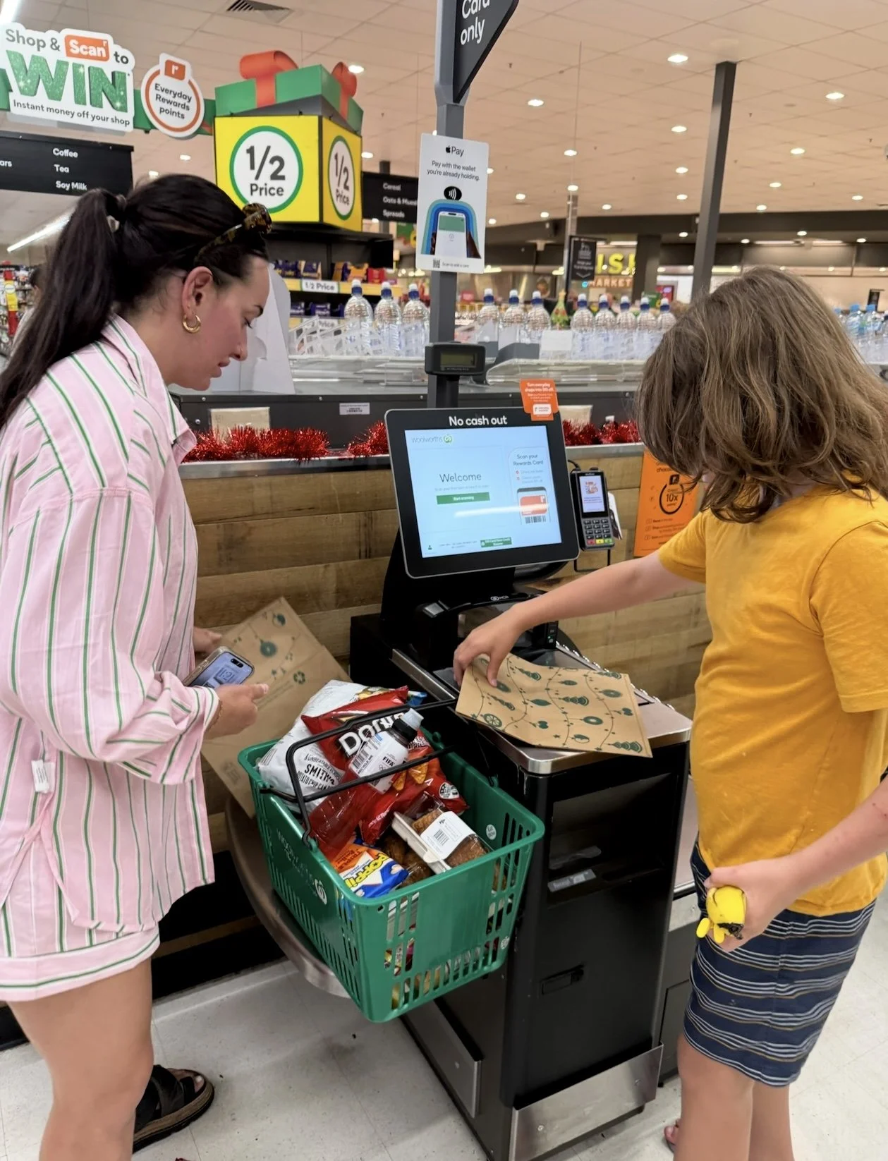 A woman and a young boy at a checkout counter in a grocery store. The woman, dressed in pink and green striped pajamas, is holding a smartphone and a paper bag. The boy, wearing a yellow t-shirt and striped shorts, is scanning items or placing them on the conveyor belt. There is a shopping basket filled with snacks and drinks. The checkout area has a digital screen, a scale, and promotional signs, with store shelves and water bottles visible in the background.