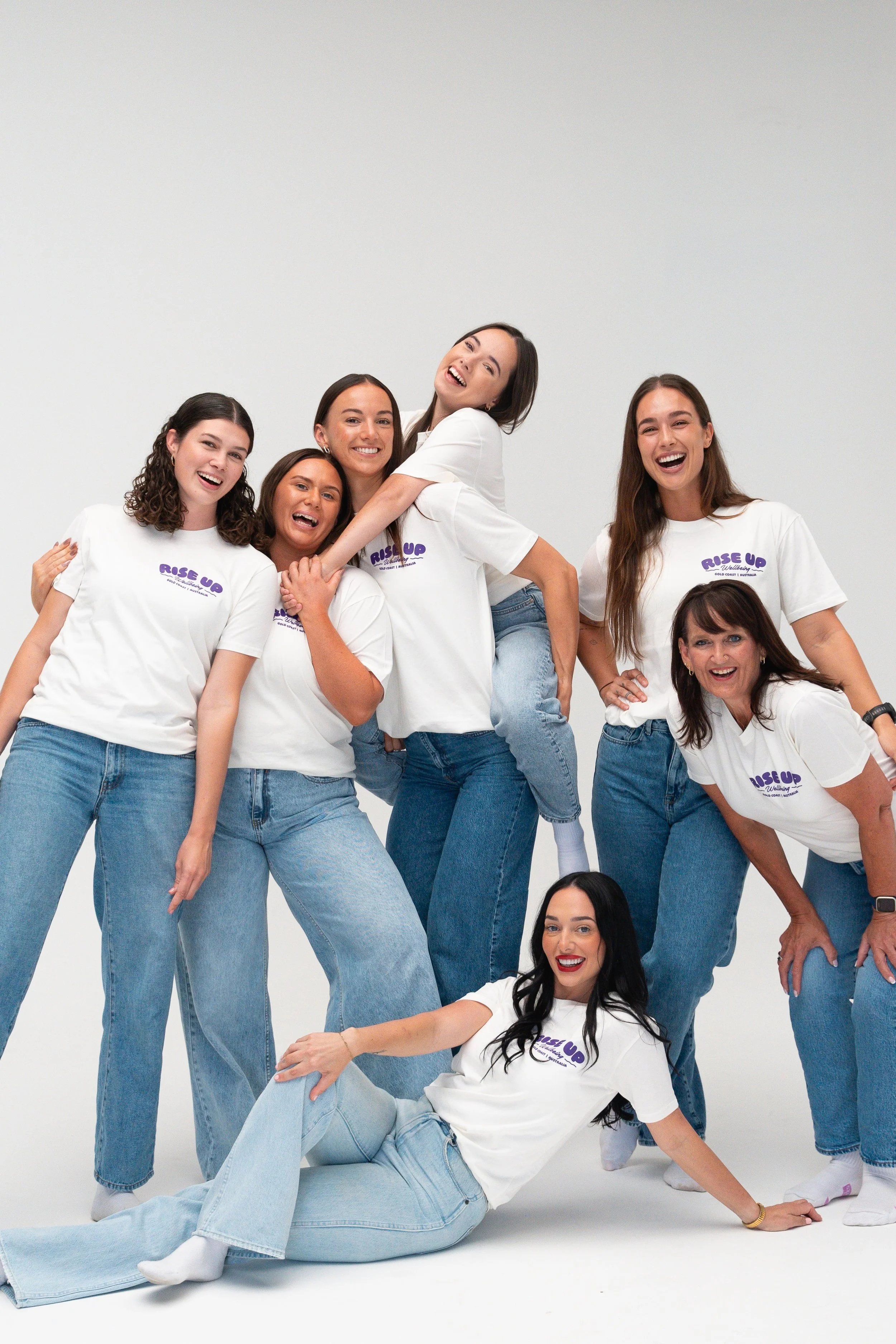 Group of six women posing and smiling, wearing white T-shirts with purple text that says 'Rise Up' and blue jeans, against a plain light background.