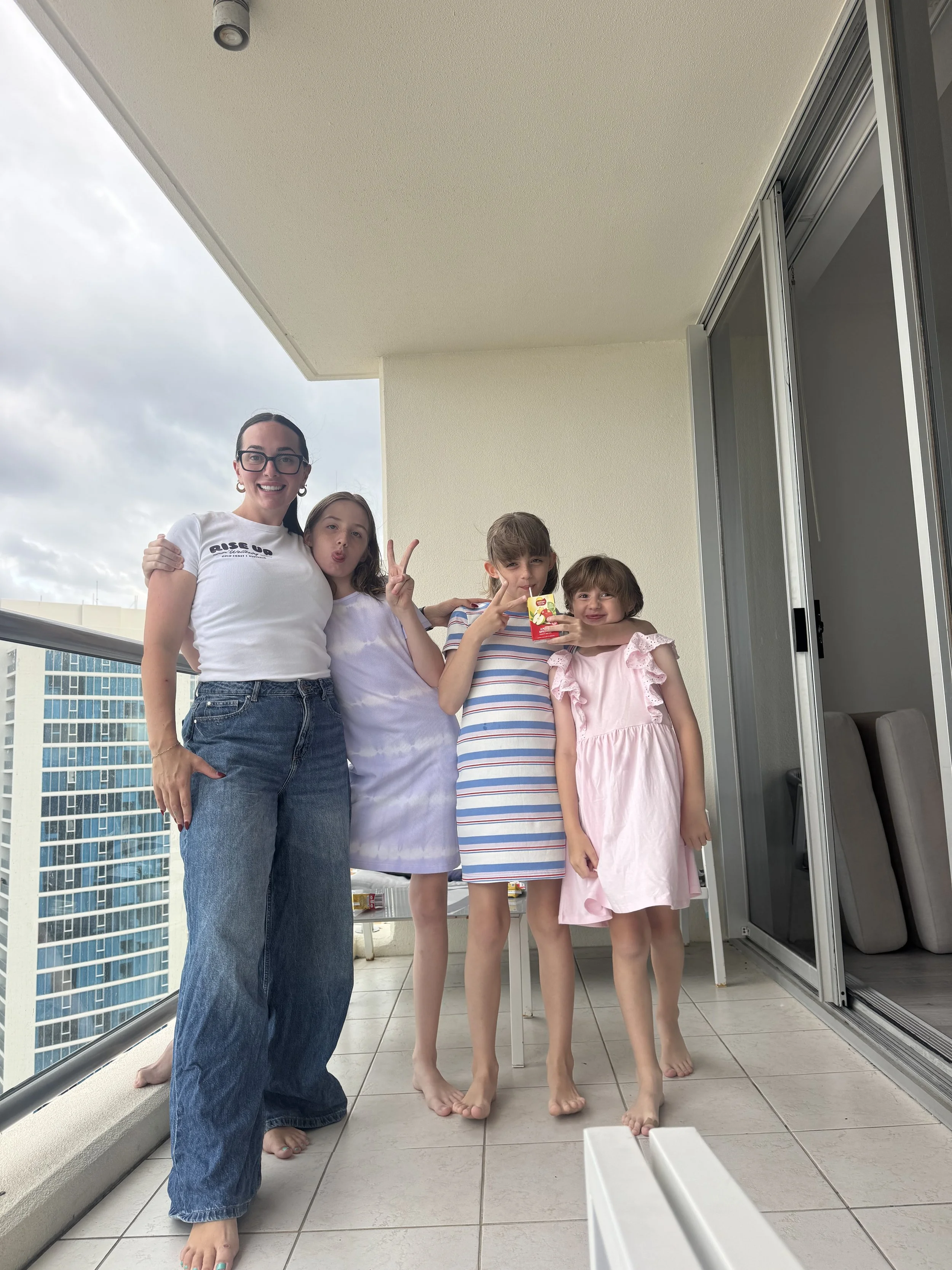 Five people, one woman and four children, on a balcony with city buildings in the background. The woman and children are smiling and making peace signs, with one child holding a juice box.