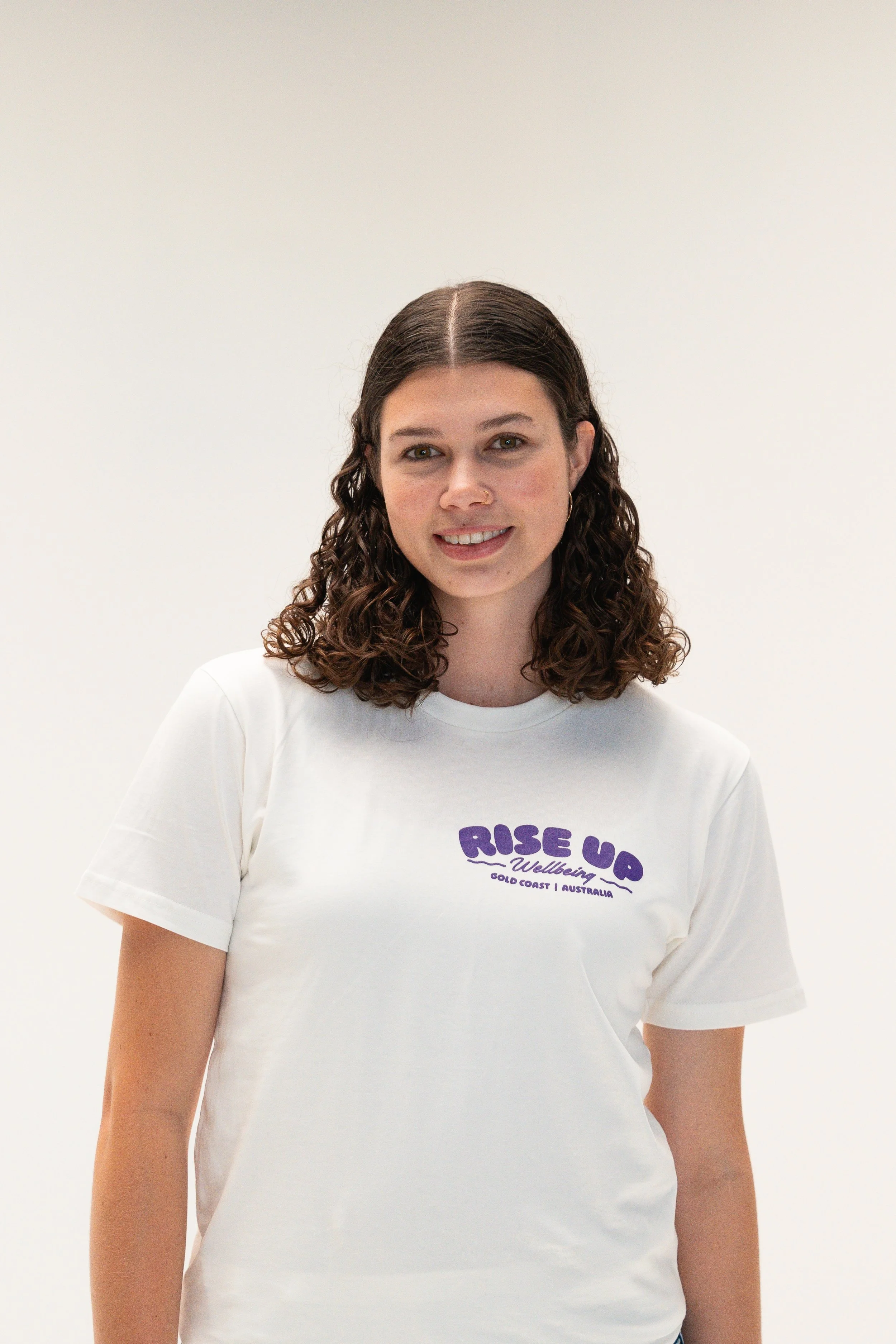 A young woman with curly brown hair, smiling, wearing a white t-shirt with purple text that reads "RISE UP Wellbeing Gold Coast | Australia," standing against a plain white background.
