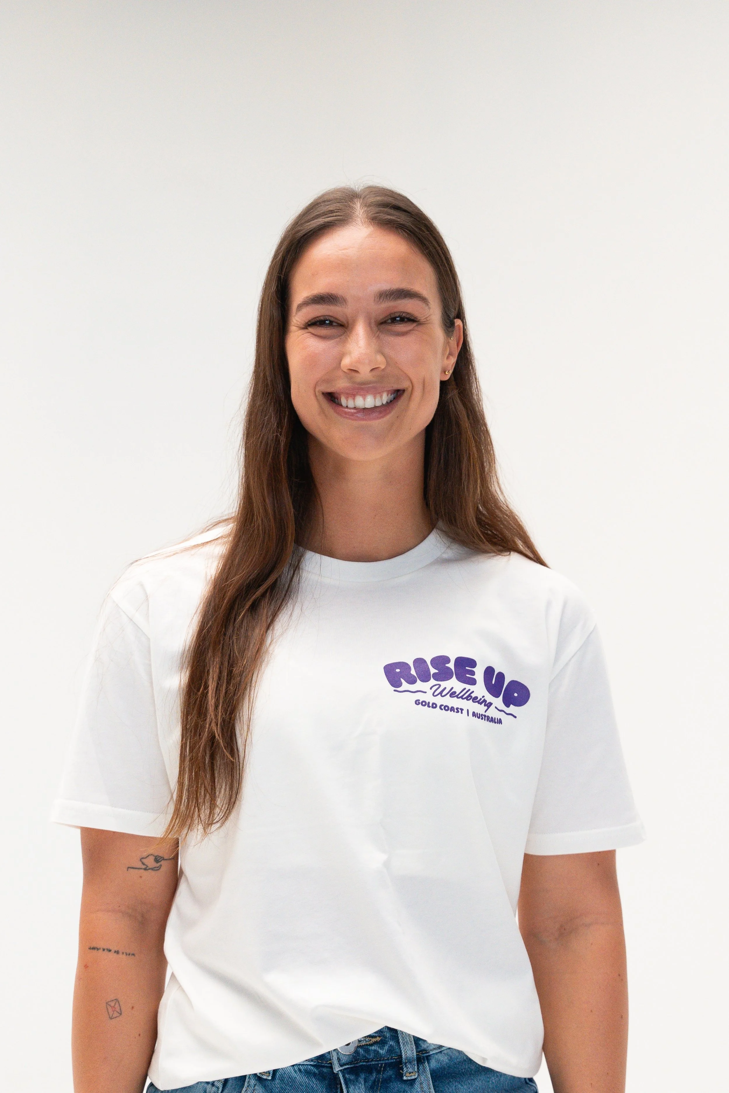 A woman with long brown hair wearing a white t-shirt with purple text that reads "Rise Up Wellbeing Gold Coast Australia." She is smiling with closed eyes and standing against a plain white background.