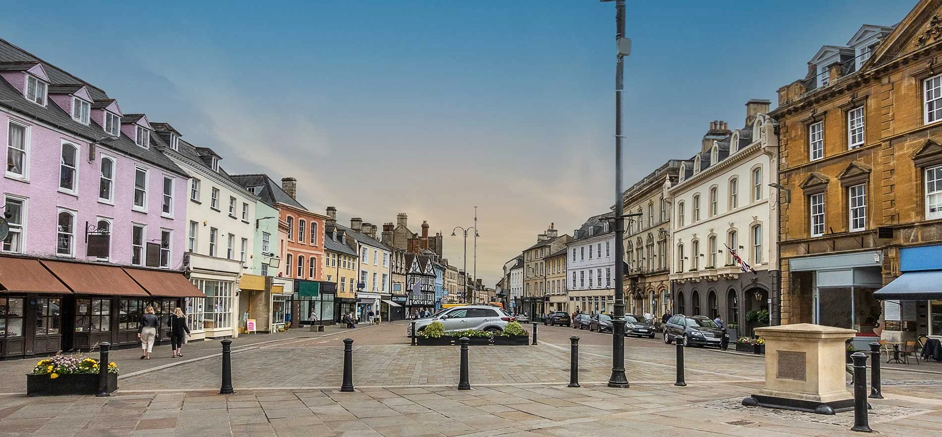 A city street with colorful buildings, parked cars, pedestrians, flower planters, and a clear sky.