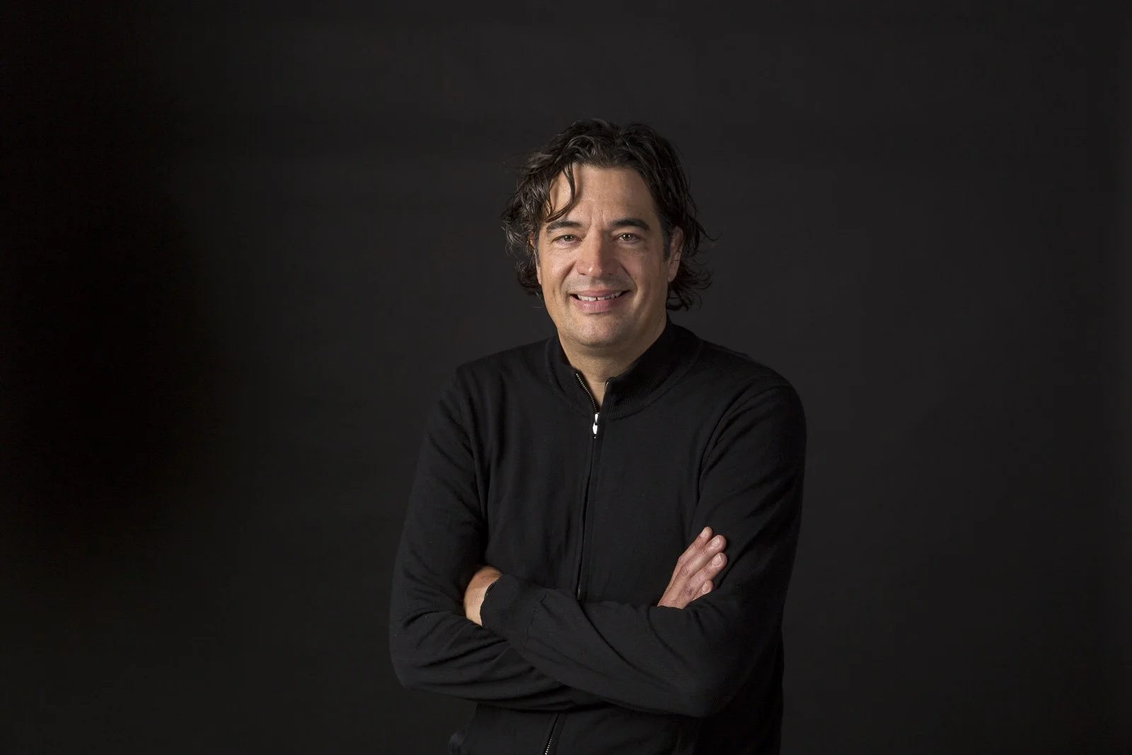 Porträt eines lächelnden Mannes mit schwarzen Haaren, der eine schwarze Jacke trägt, vor dunklem Hintergrund.