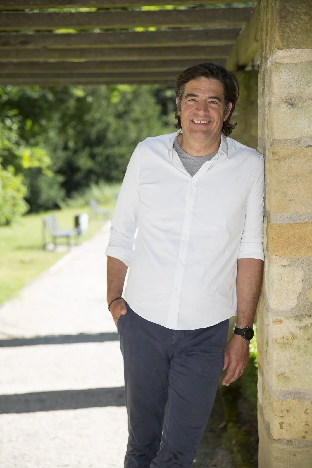 Ein lächelnder Mann in weißem Hemd und dunklen Hosen steht draußen an einer Steinmauer unter einem Holzüberhang, im Hintergrund grüne Bäume und eine Bank.